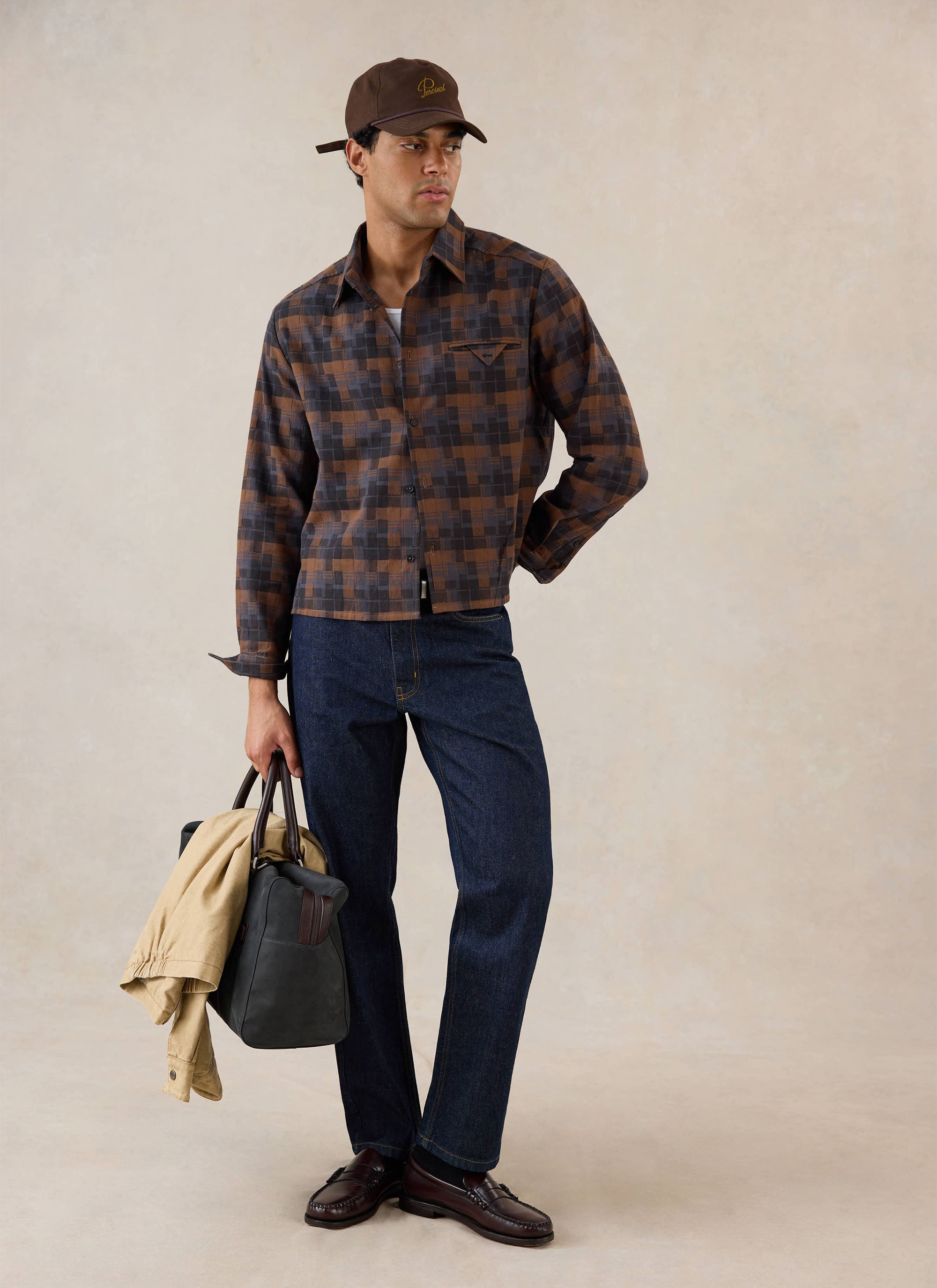 A model wears the brown check Hinsley boxy long sleeve shirt from Percival, styled with dark jeans, a cap, and holding a bag.