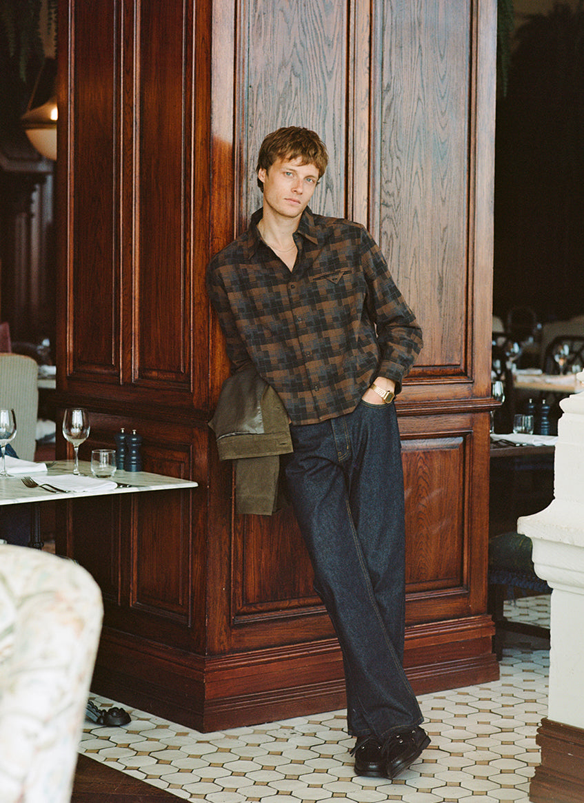 A model leans against a wooden column wearing the Percival brown check Hinsley boxy long sleeve shirt with raw jeans.