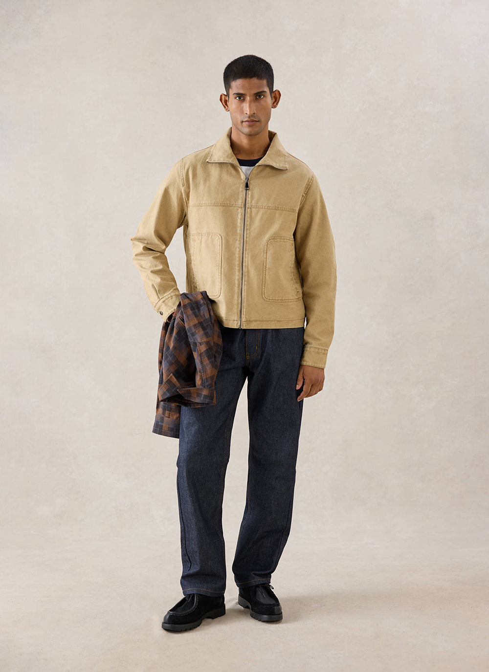 A model wears the cream Percival Jackson Harrington Jacket, holding a checked shirt with dark jeans in a studio.
