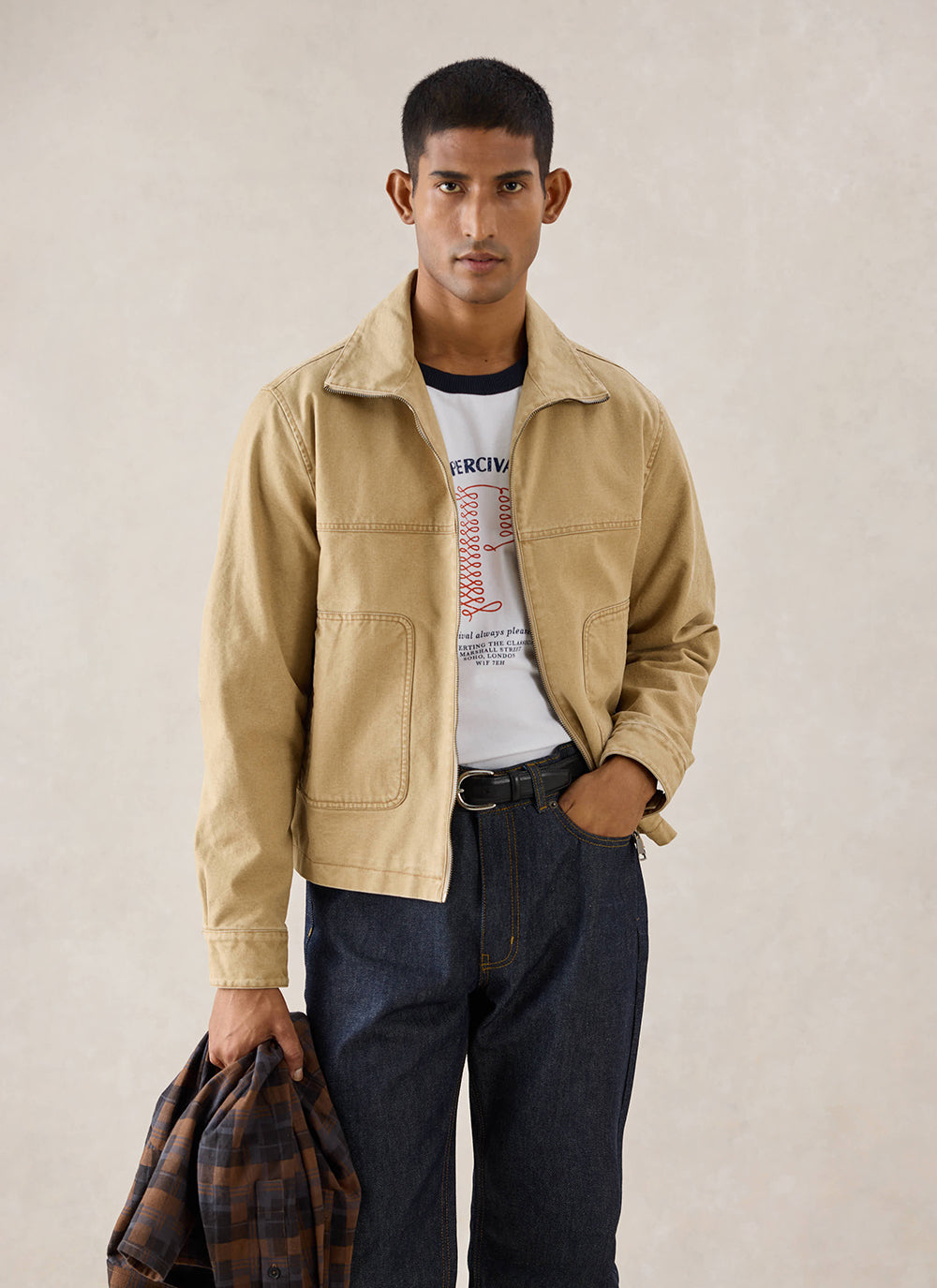 A model wears the cream Percival Harrington Jacket, styled with a t-shirt, dark jeans, and holding a patterned shirt.