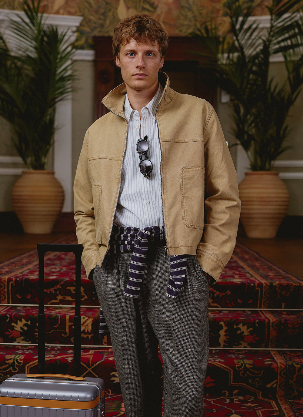 A model wears the beige Percival Jackson jacket over a striped shirt and patterned trousers in a hotel interior.