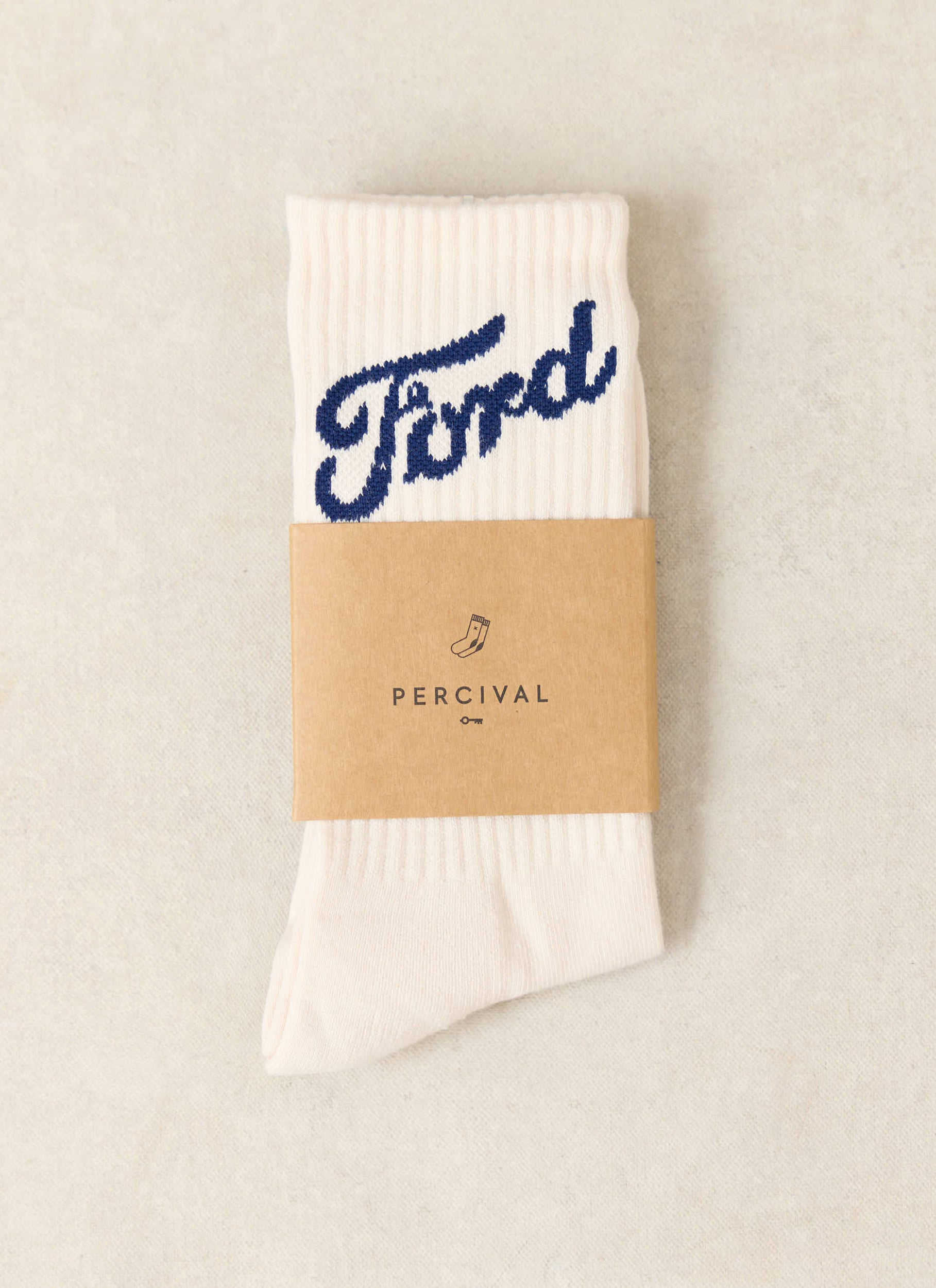 Cream Ford x Percival jacquard crew socks with a blue logo, presented in a brown paper branded sleeve.