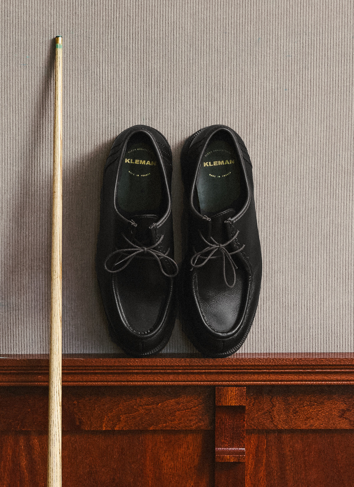 A pair of black Percival Kleman Padror shoes placed against a textured wall next to a cue stick.