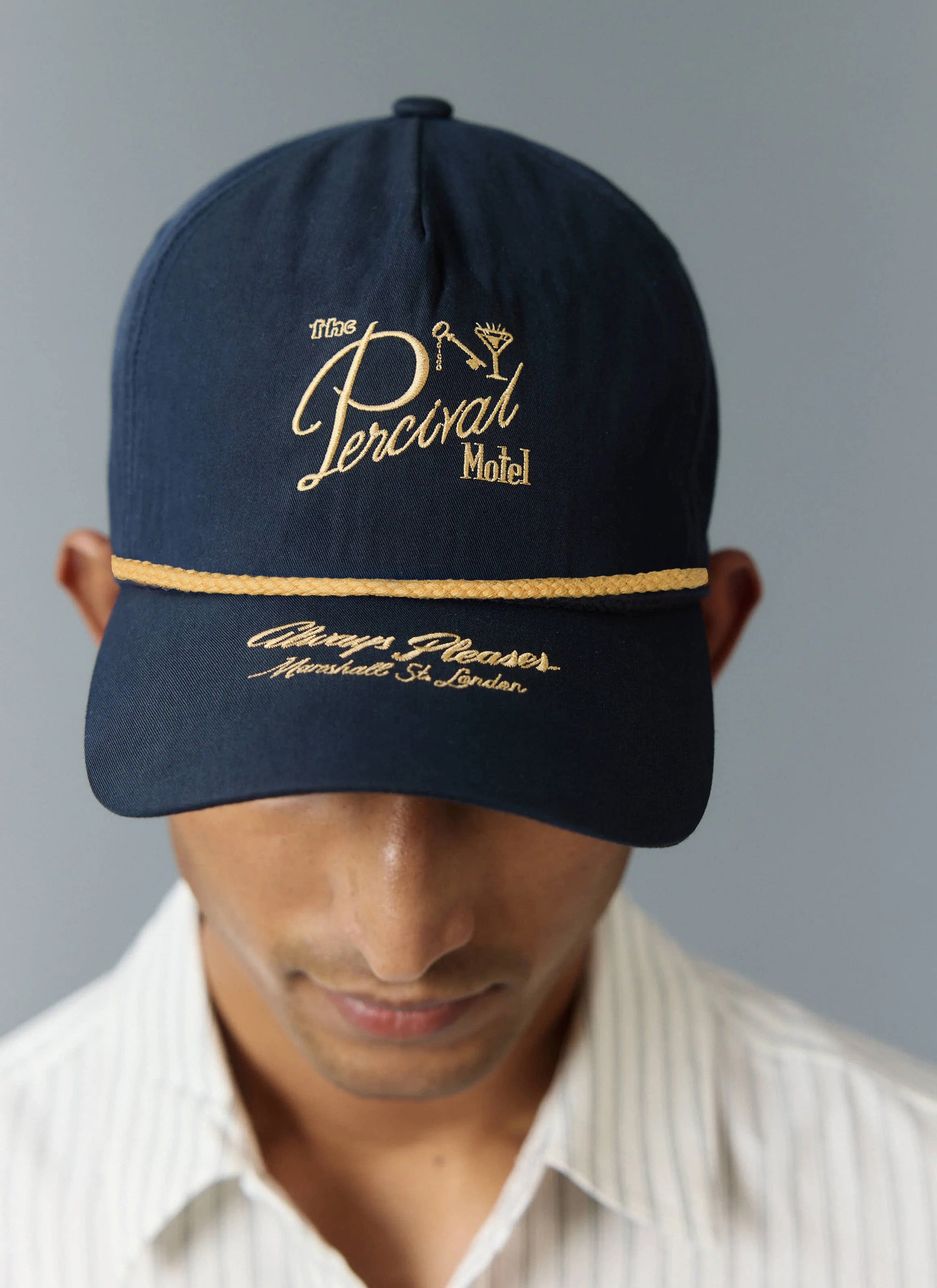 Percival Kingsley navy cotton soft trucker cap with gold motel embroidery and cord detail, worn by a model.