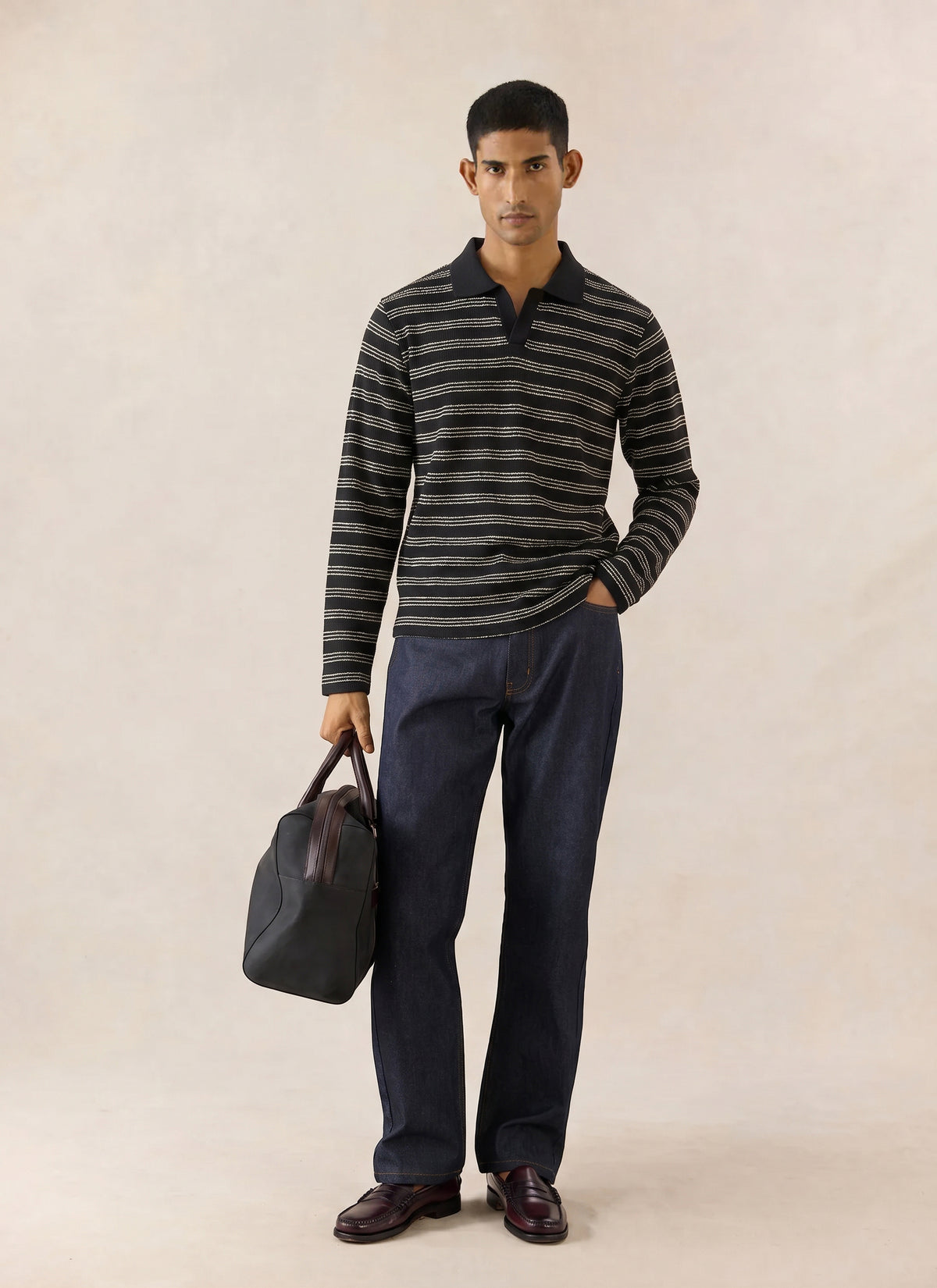 A model wears the black striped Percival Laurent Long Sleeve Polo with dark denim jeans and holds a holdall bag.