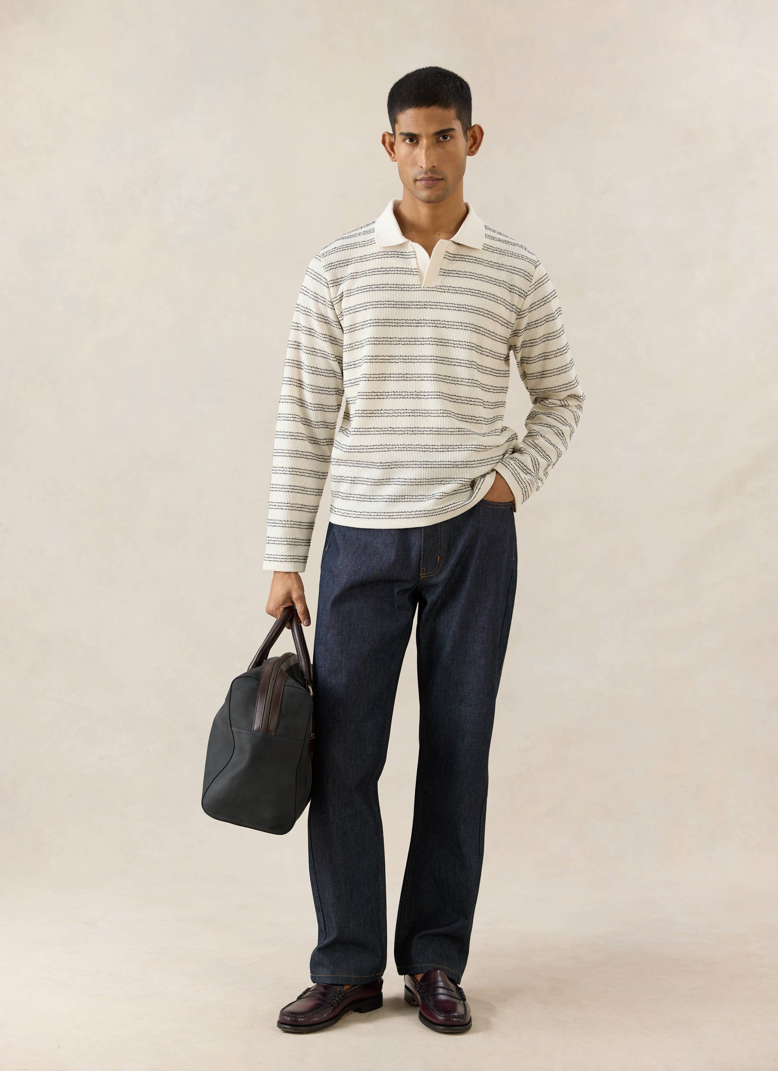 The Percival Laurent cream long sleeve polo with stripes, worn by a model with raw indigo denim jeans and a travel bag.
