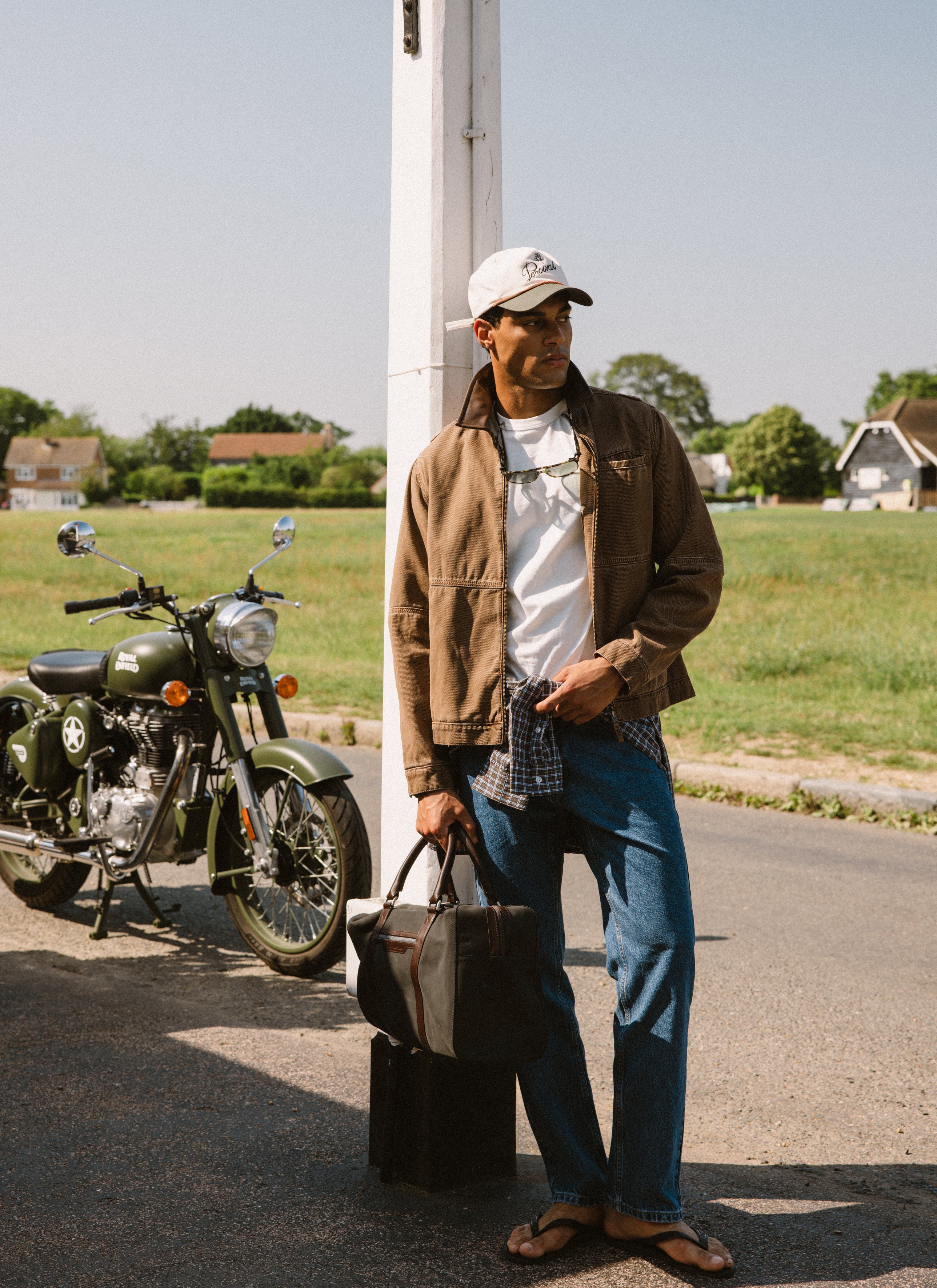 A model wears the espresso Percival Lucas Jacket, styled with a cap and jeans by a green motorcycle.