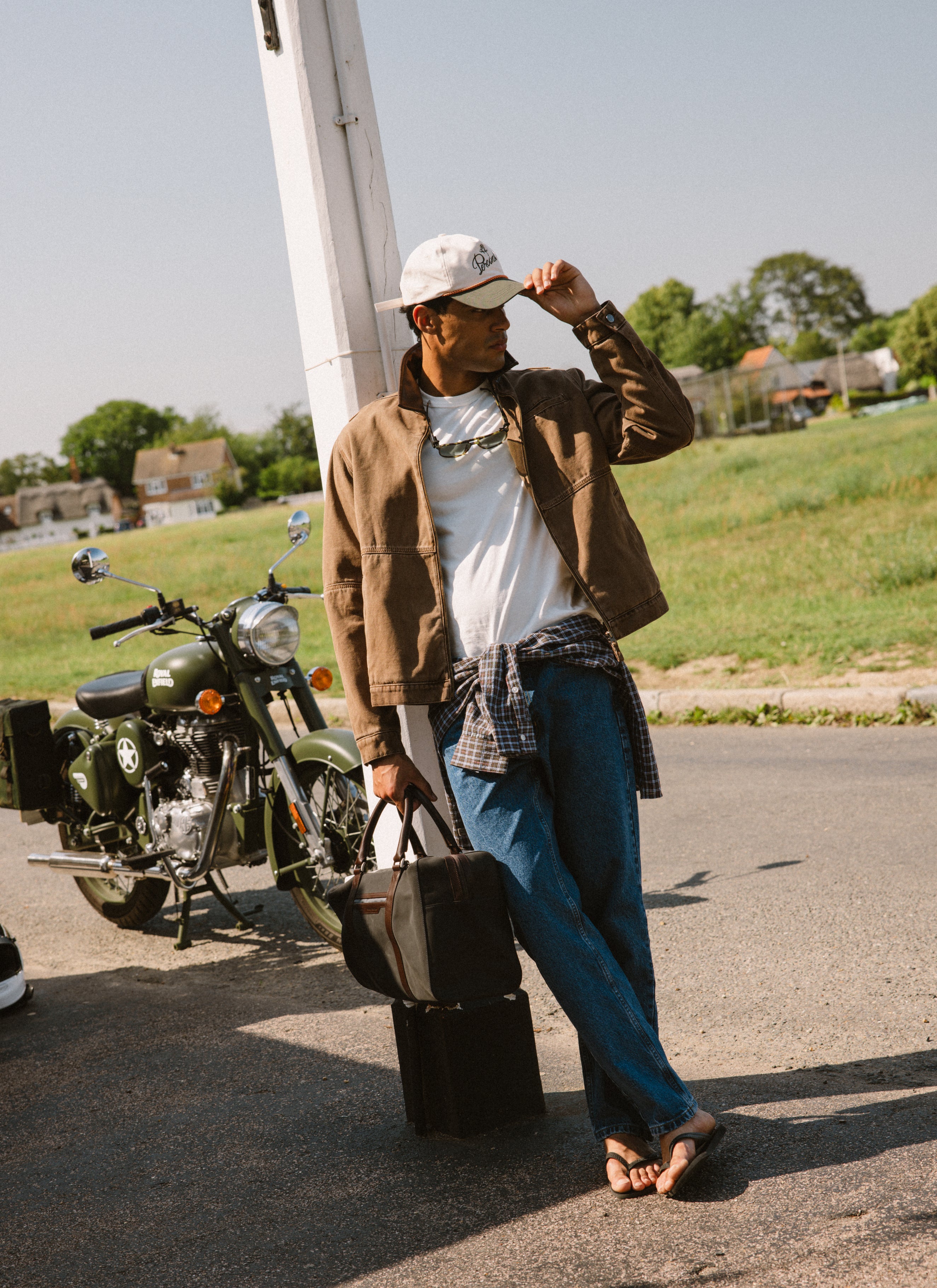 A model wears the mocha brown Percival cotton twill jacket next to a motorcycle outdoors.