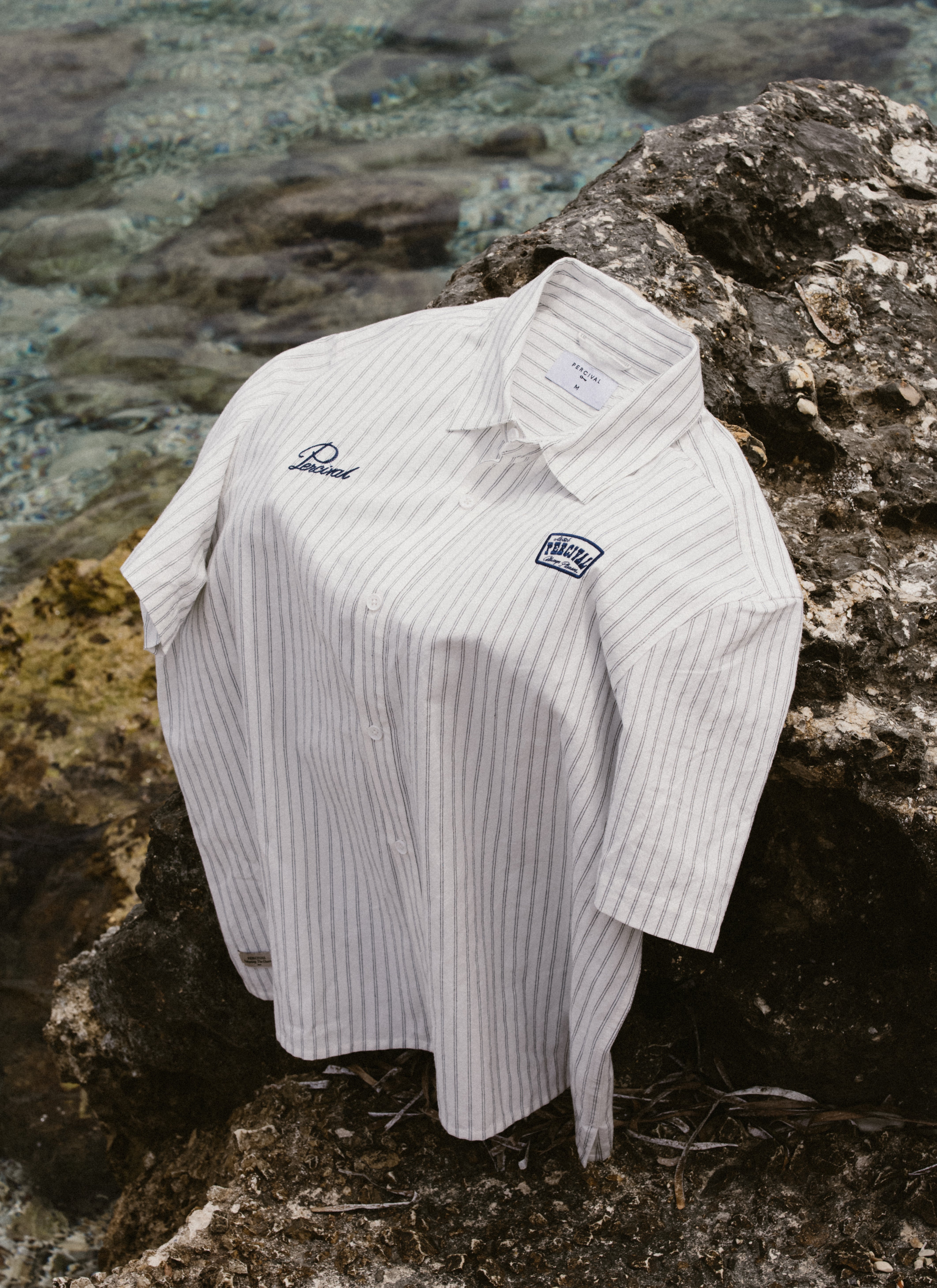 Percival Lucia short-sleeve shirt in cream stripe cotton with a motel badge, draped over rugged rocks by the sea.