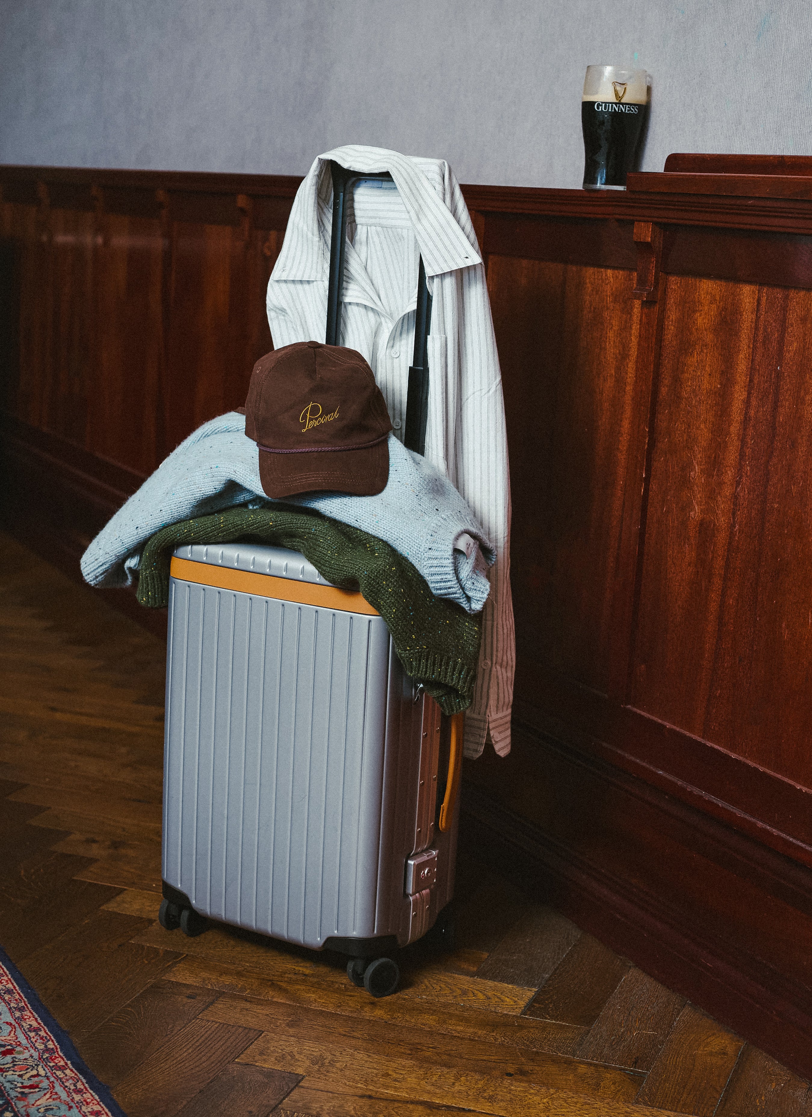 A silver suitcase packed with Percival clothing and a cap, standing against a wood-panelled wall with a pint glass.