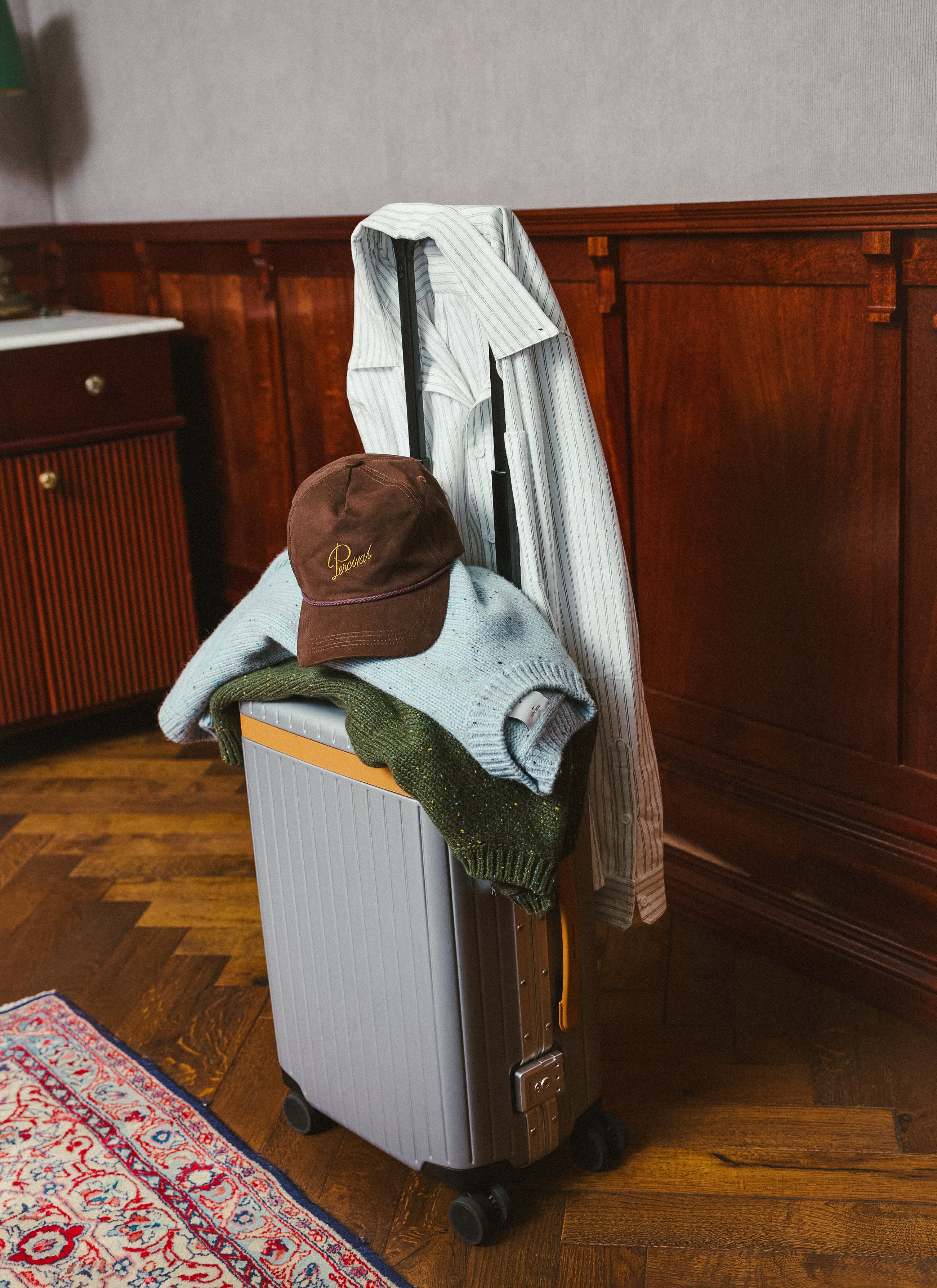 A suitcase packed with Percival clothing including a white striped shirt, brown cap, and jumpers.