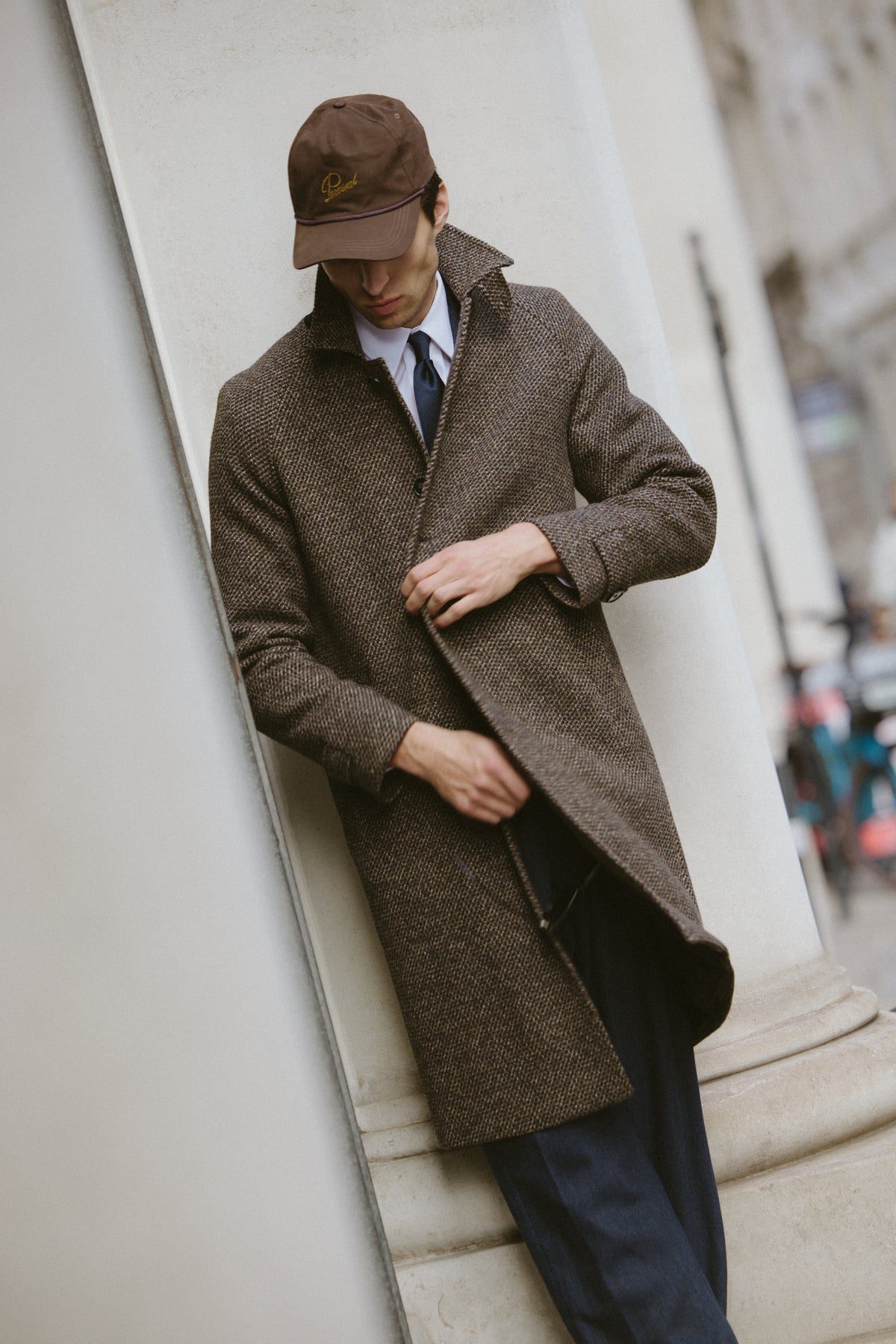 A model wearing the brown fleck Percival wool Mason car coat and a matching cap while leaning against a column.