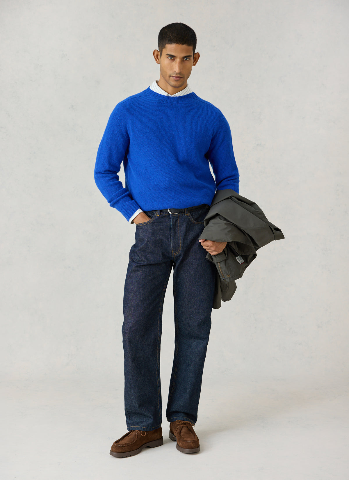 A model wearing Percival's cobalt blue lambswool jumper with a collared shirt, dark jeans, and holding a grey jacket.