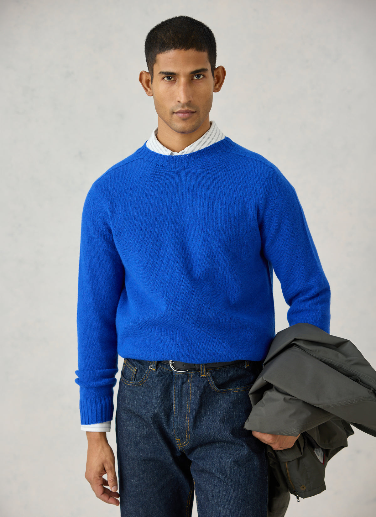 A model wearing Percival's cobalt blue lambswool jumper over a striped shirt, holding a dark jacket.