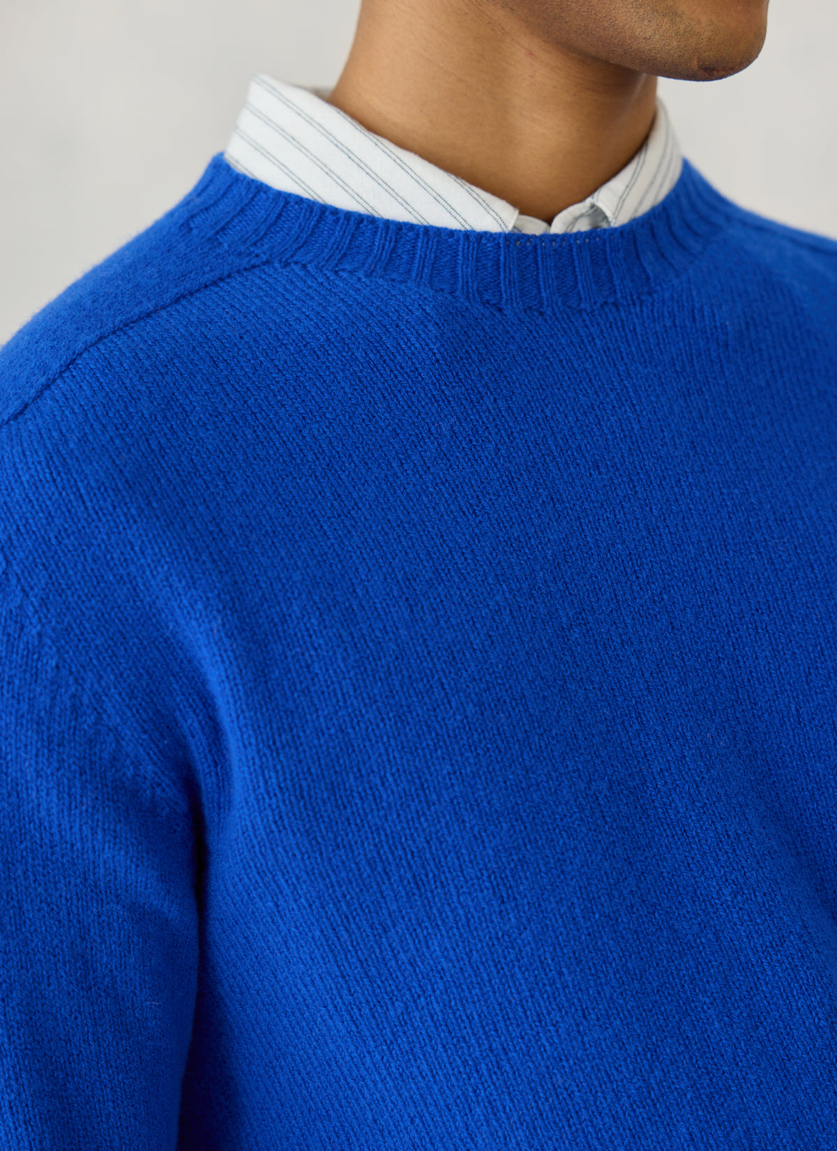 A close-up of the Percival cobalt blue lambswool jumper showing the ribbed crew neck layered over a striped shirt.