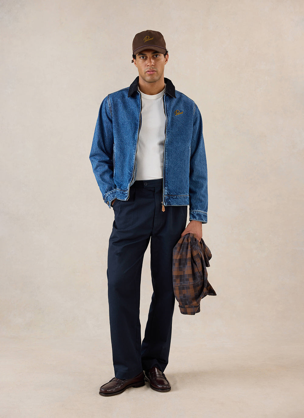 A model wears the mid wash blue Percival Mackey Zip Jacket, styled with a white tee, dark trousers and a brown cap.