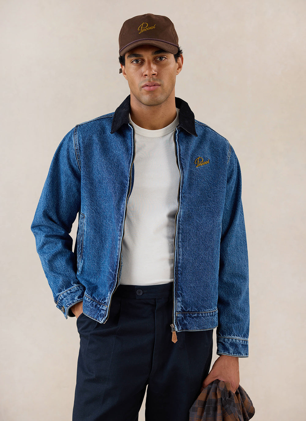 A model wears the mid wash blue Percival Mackey Zip Jacket, styled with a brown cap and dark trousers.