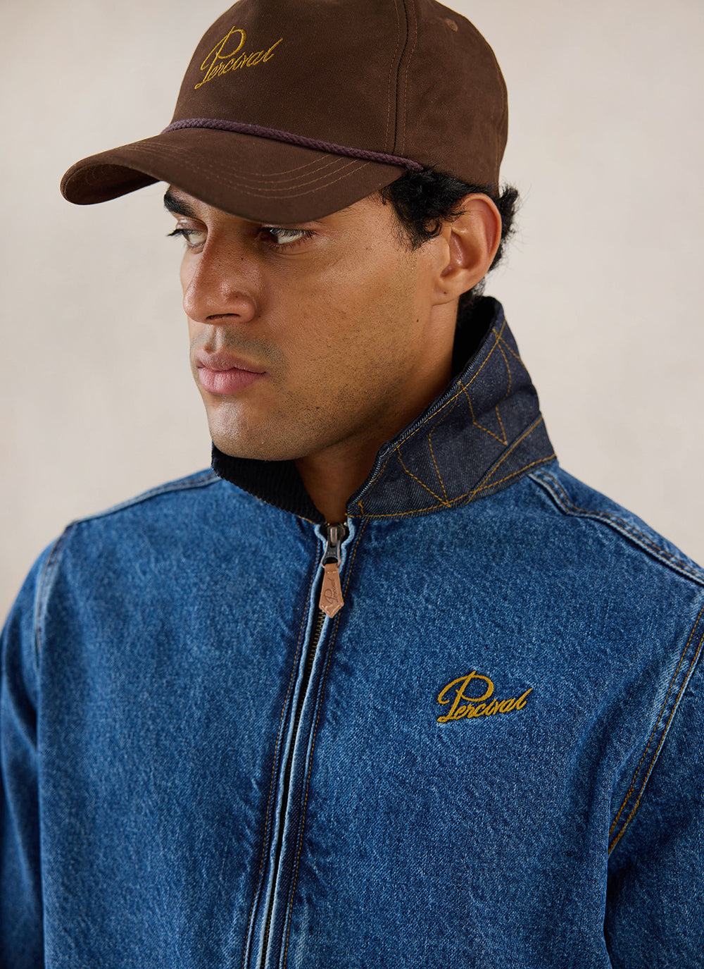 A close-up shot of a model wearing the mid wash blue Percival Mackey Embroidered Zip Jacket and a brown cap.