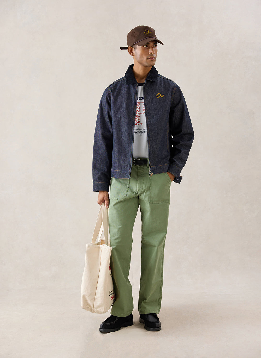 A model wears the raw denim Percival Mackey Zip Jacket, styled with green trousers, a cap, and a tote bag in a studio.