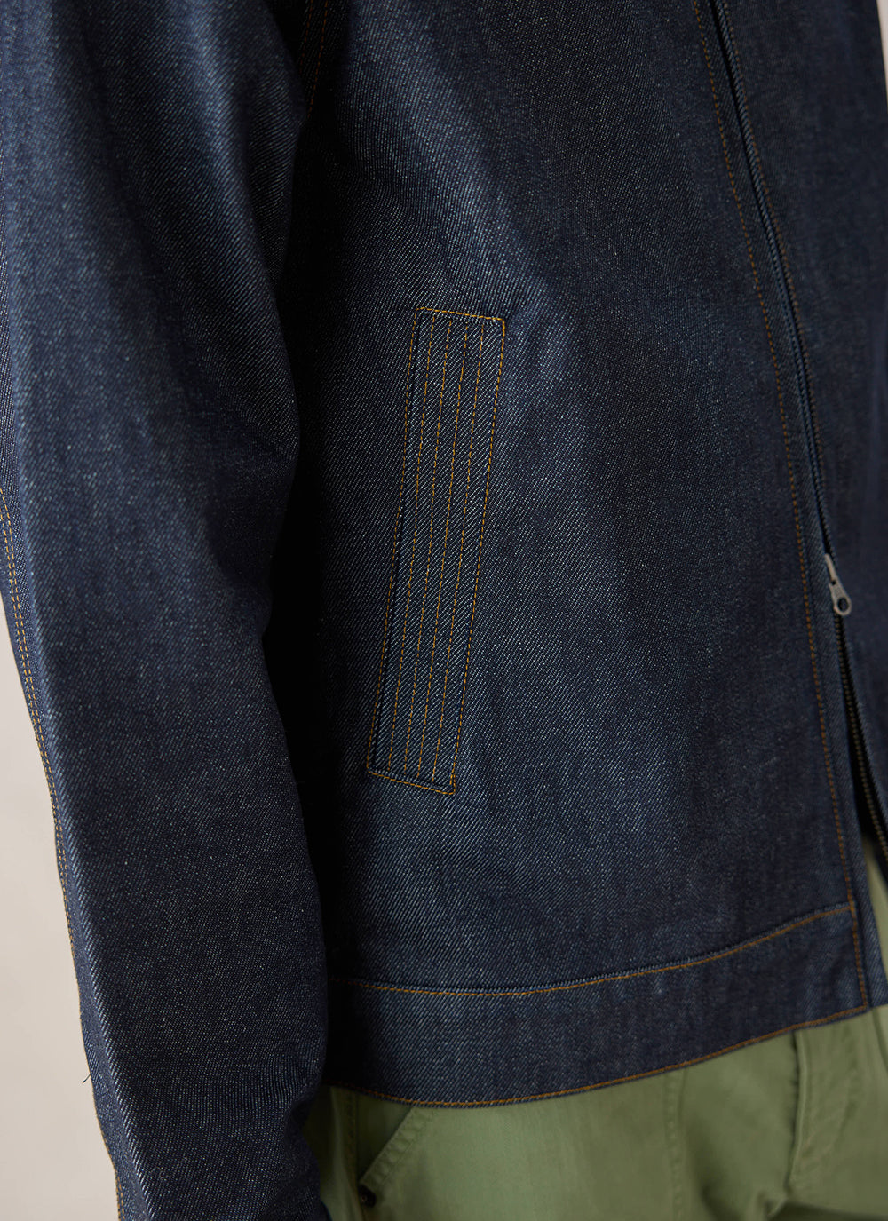 A close-up of the raw denim Mackey Zip Jacket from Percival, highlighting the pocket and contrast stitching details.