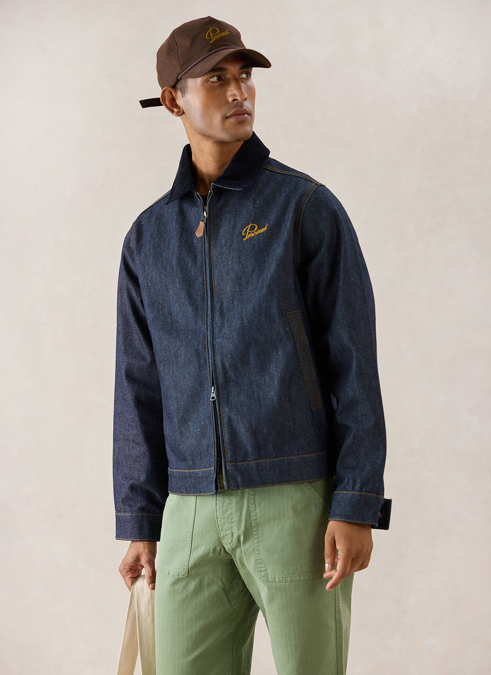 A model wears the raw denim Percival Mackey Zip Jacket, styled with a brown cap and green trousers in a studio setting.