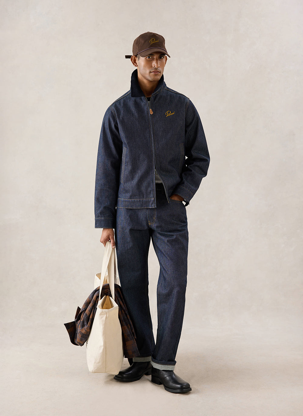 A model wears the raw denim Percival Mackey Zip Jacket, matching jeans, and a cap, holding a tote bag.