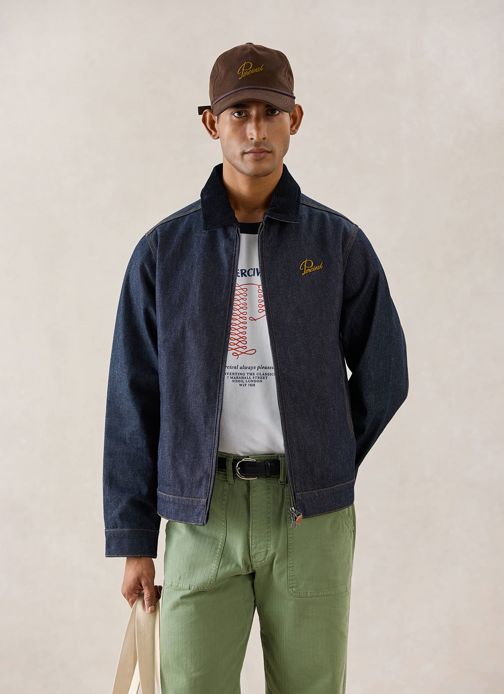A model wears the raw indigo Percival Mackey Zip Jacket, styled with a graphic t-shirt and green trousers.