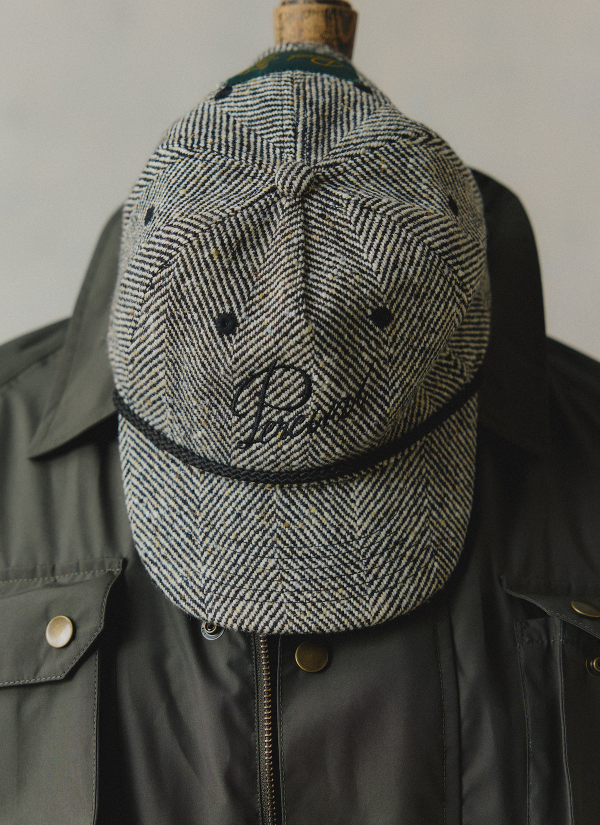 Grey herringbone wool Percival trucker cap featuring black rope trim and script embroidery, displayed on a jacket.