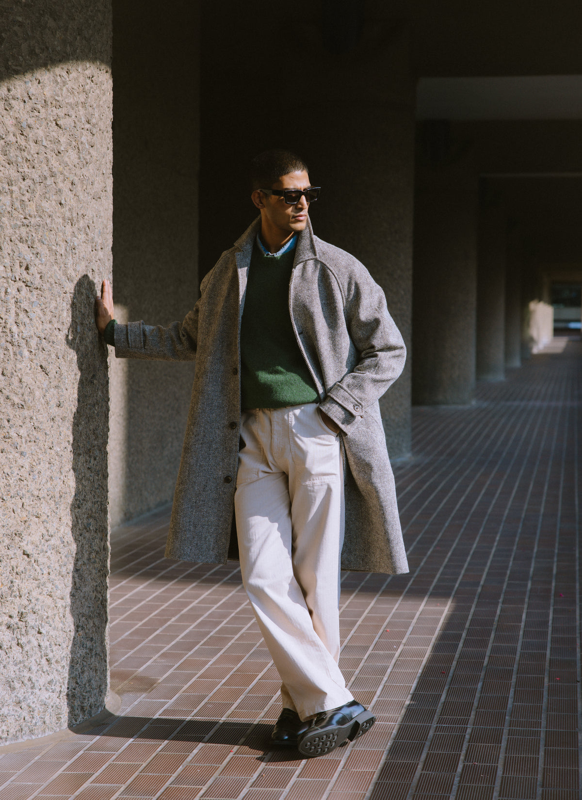 A model wearing the grey herringbone wool Percival car coat, green lambswool jumper, and black leather shoes outdoors.
