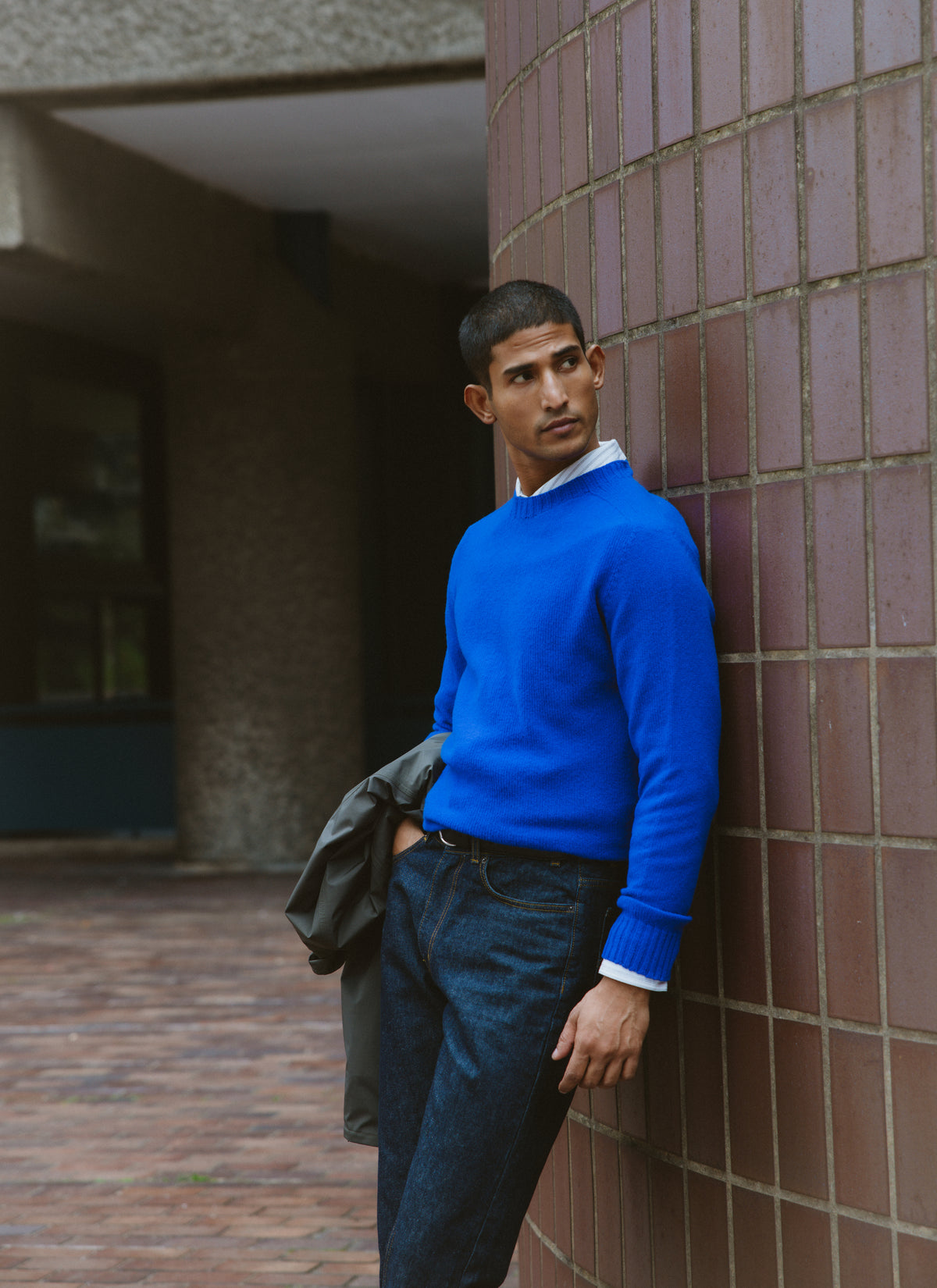 A model wearing Percival's cobalt blue lambswool jumper and dark jeans, leaning against a tiled column outdoors.