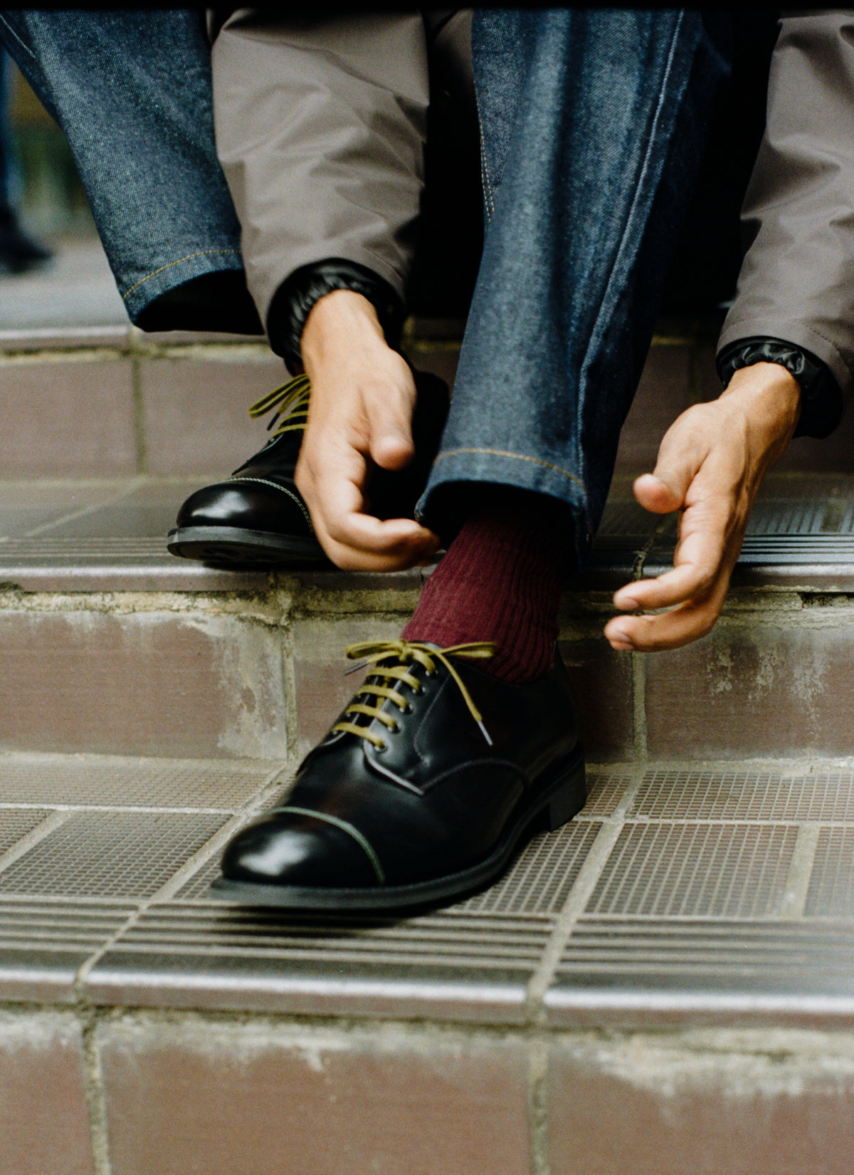 A model wearing Percival's black leather Derby shoes with green laces and stitching, styled with denim jeans on stone steps.