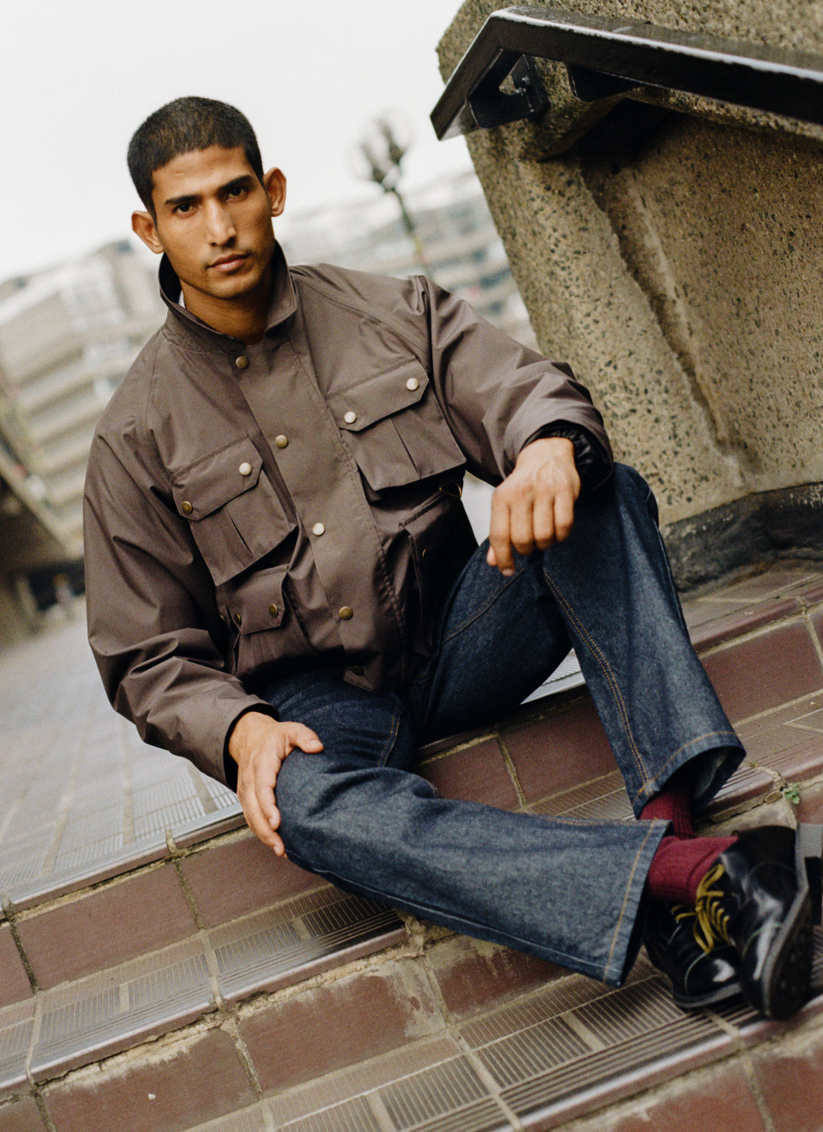 A model sitting outdoors on stone steps wearing a brown hunter jacket, dark jeans, and black leather Derby shoes.