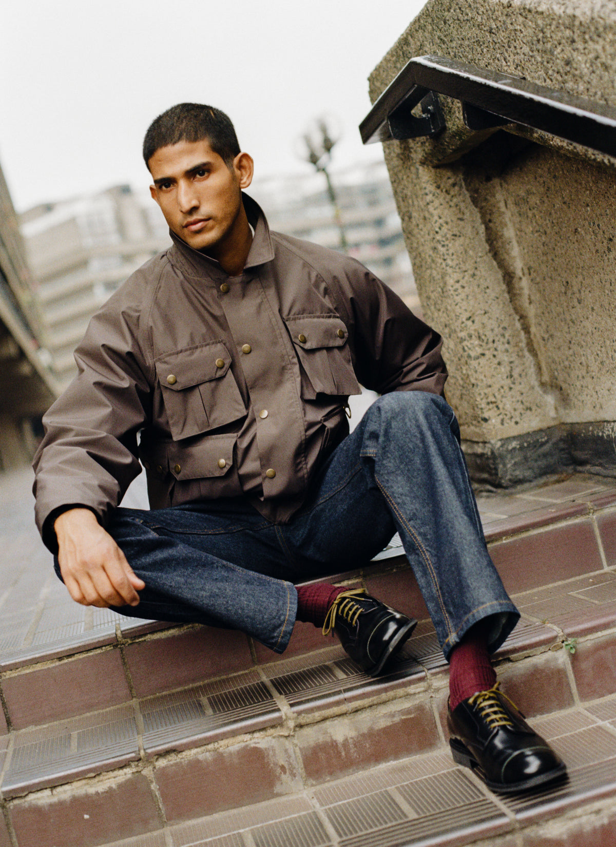 A model sitting outdoors on stone steps wearing the Percival black leather Derby shoes, jeans, and a brown hunter jacket.