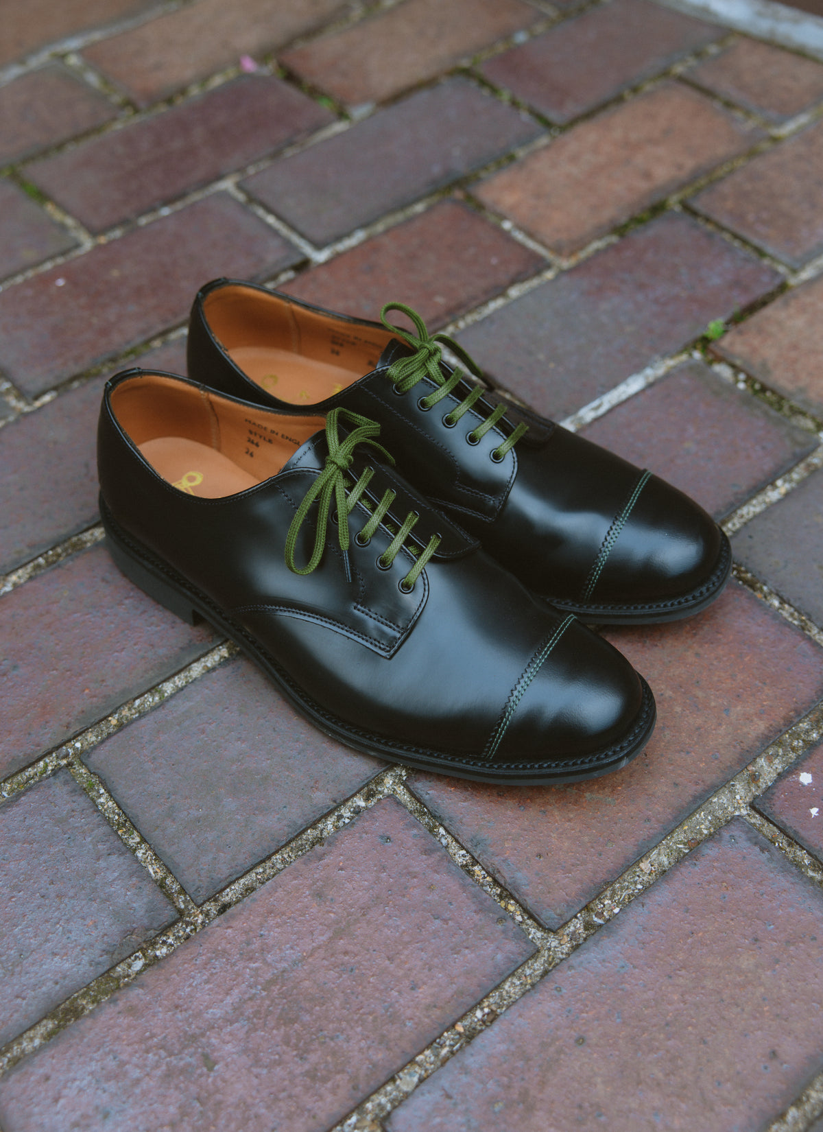 Black leather Derby shoes made in England with military green laces and stitching, displayed on a brick surface.