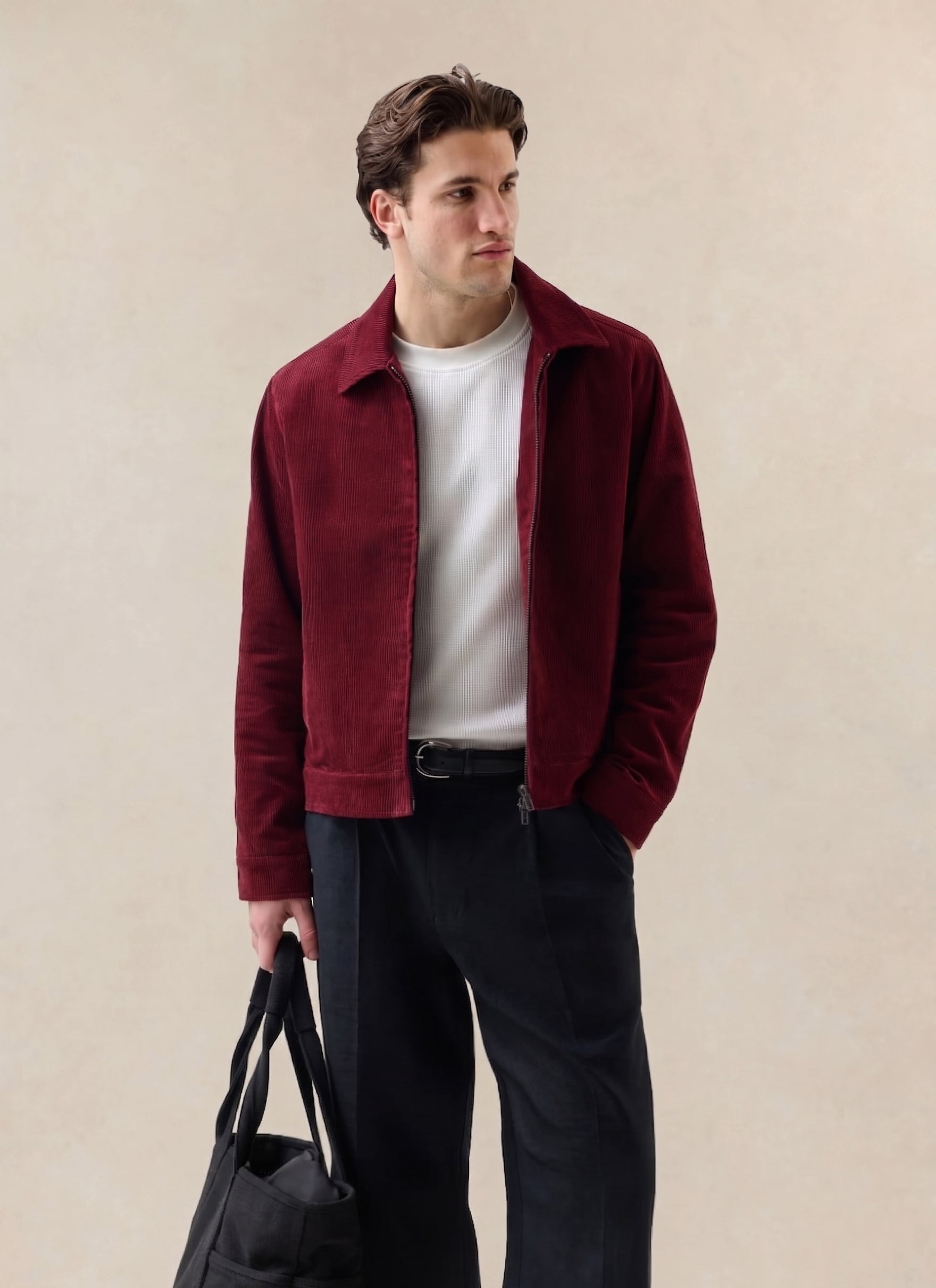 A model wears the burgundy Marcel cord jacket from Percival over a white t-shirt, styled with dark trousers and a black bag.