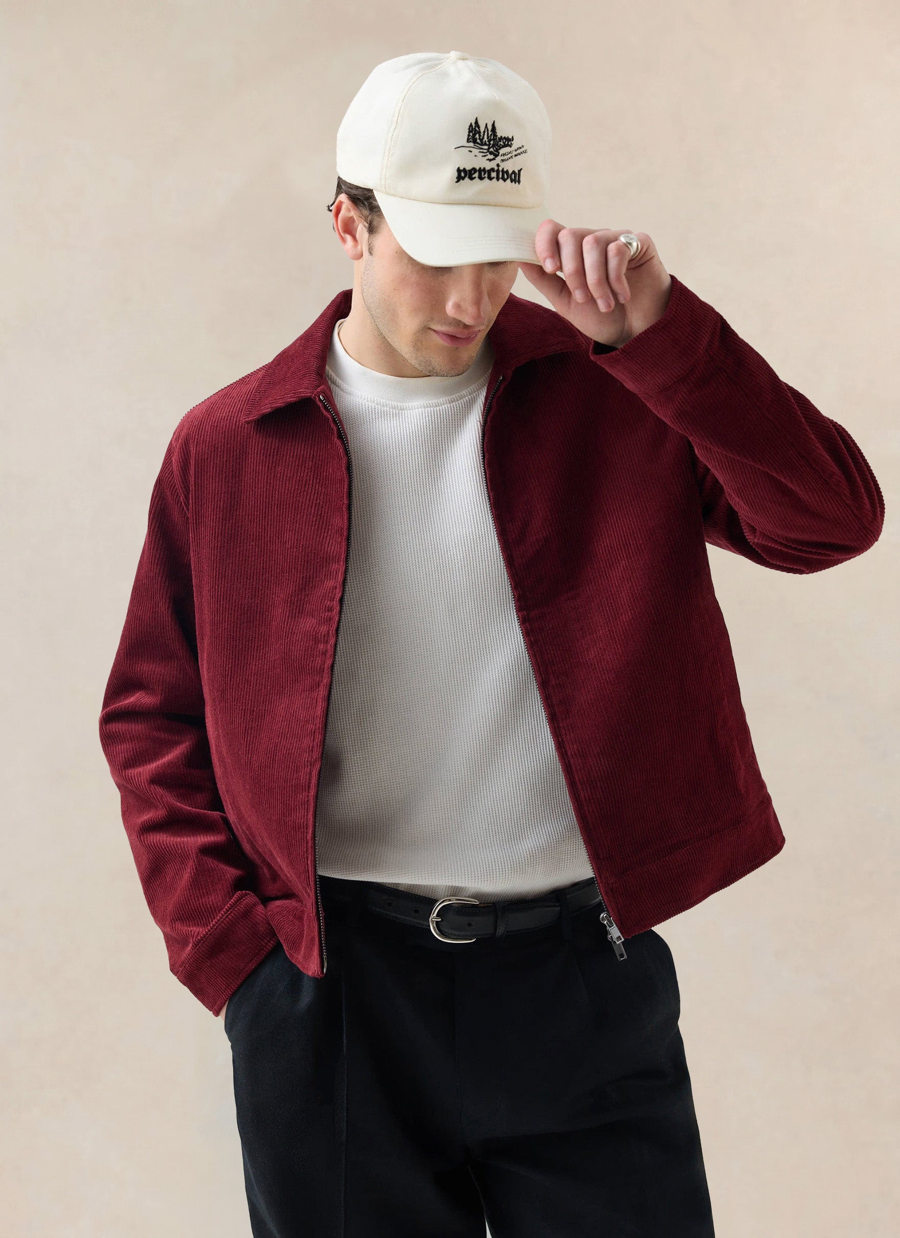 A model wears the burgundy Marcel cord jacket from Percival over a white t-shirt and black trousers, adjusting his cream cap.