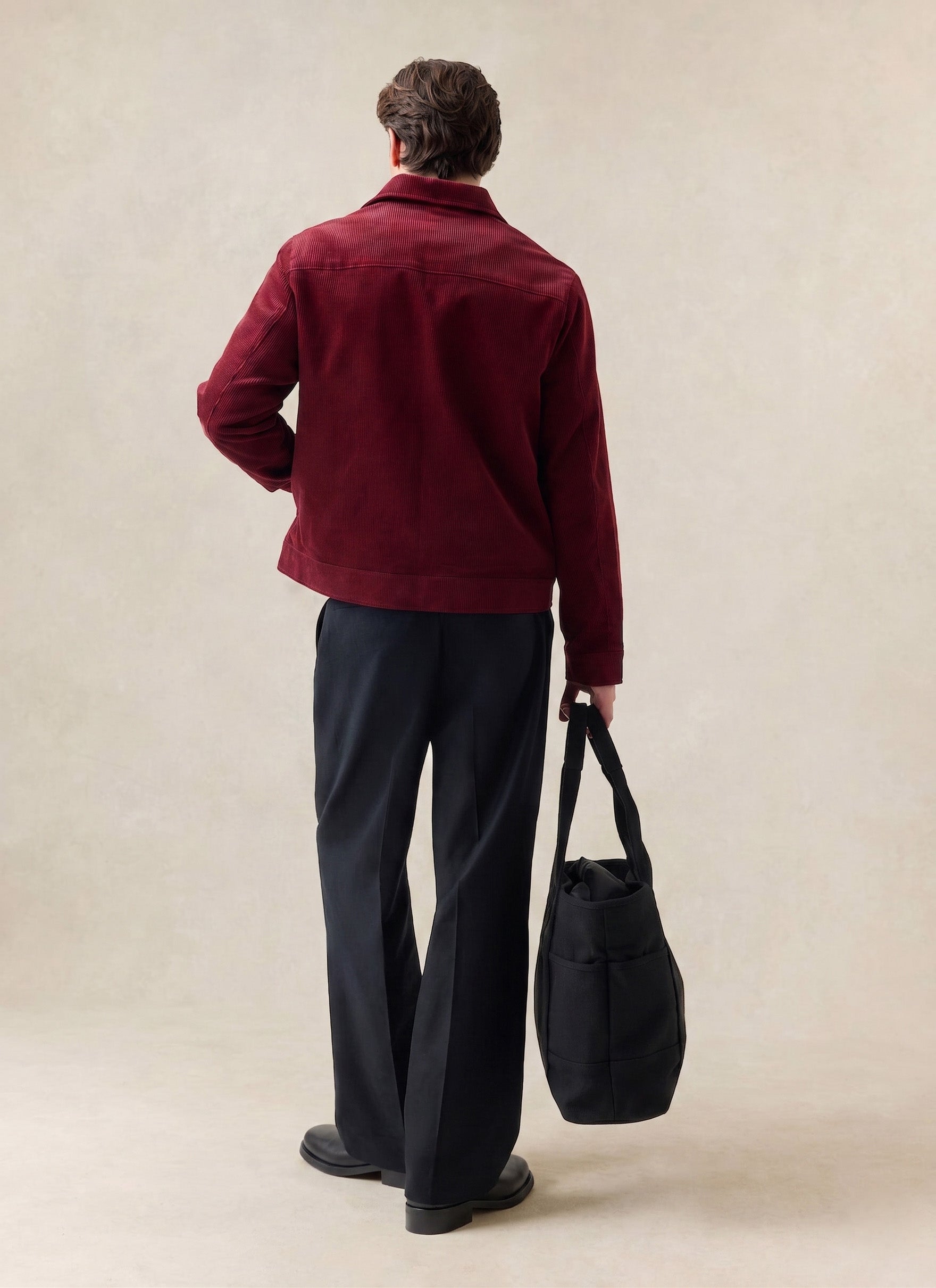 A rear-view shot of a model wearing the burgundy Marcel Cord Jacket from Percival, styled with dark trousers and a black bag.