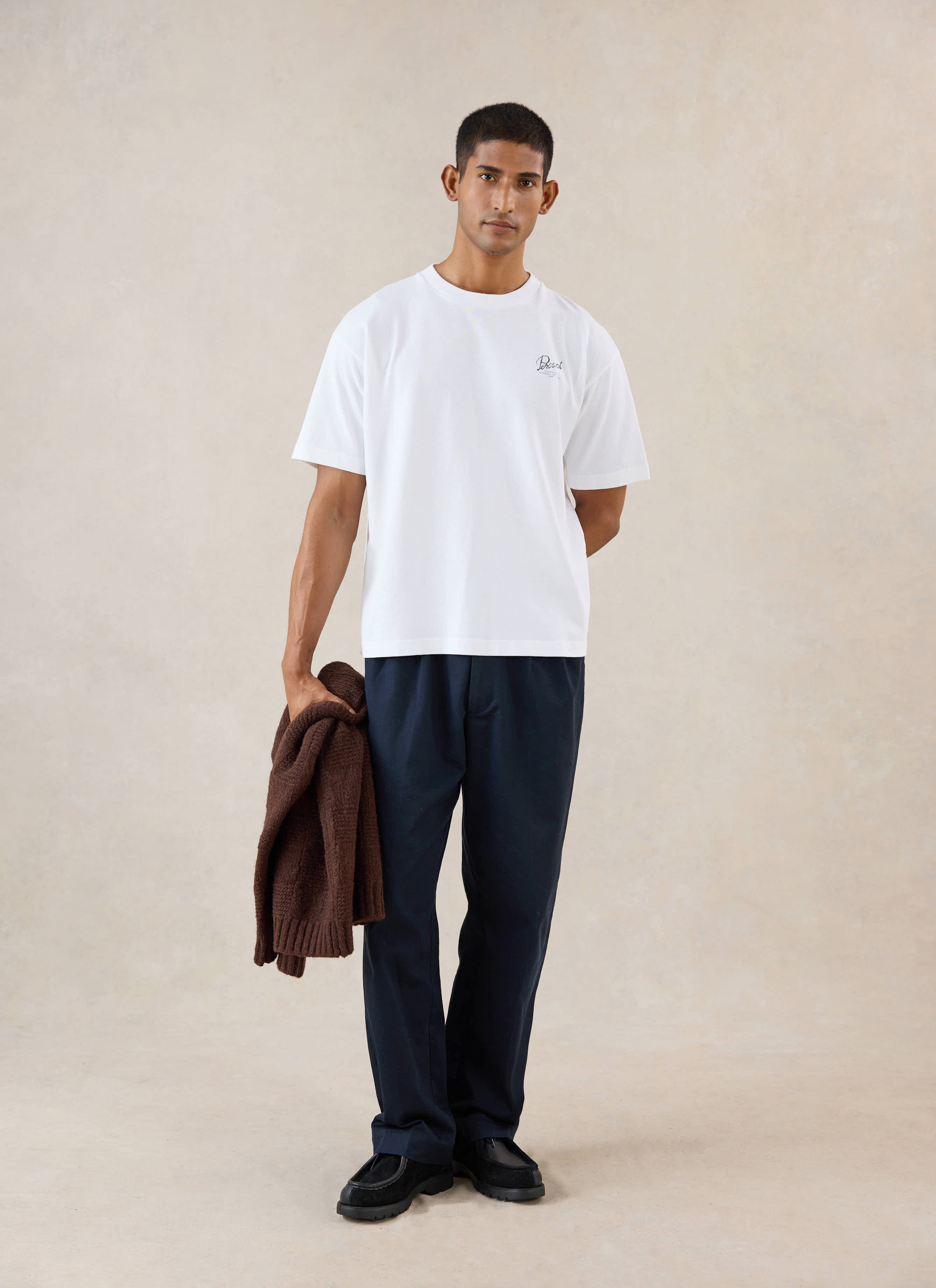 A model wears the white Percival Marley boxy t-shirt, styled with dark trousers and holding a brown knit jumper in a studio.