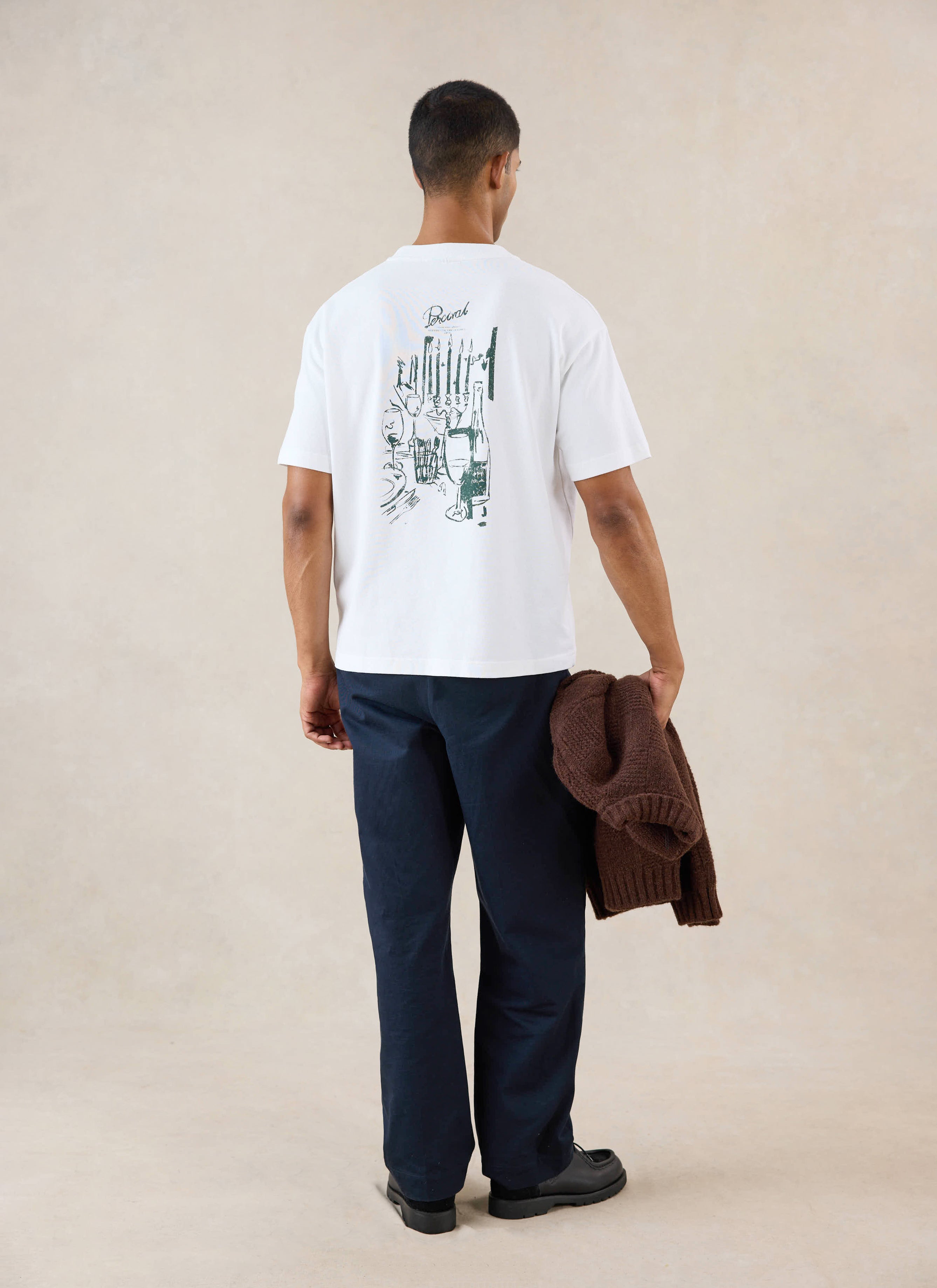 A model from behind wears the white Percival Marley boxy T-shirt featuring a dinner party back graphic and dark trousers.