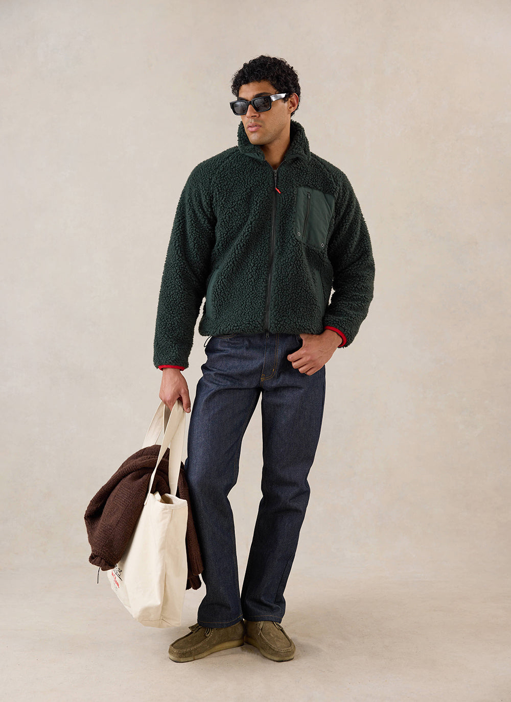 A model wears the forest Percival fleece jacket, styled with dark denim jeans and holding a tote bag in a studio.