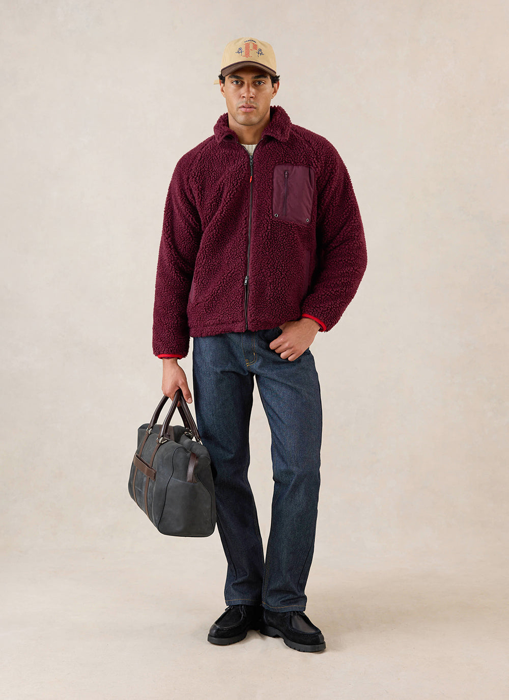 A model wears the merlot Percival wool fleece jacket, styled with dark jeans, a cap, and a holdall bag.
