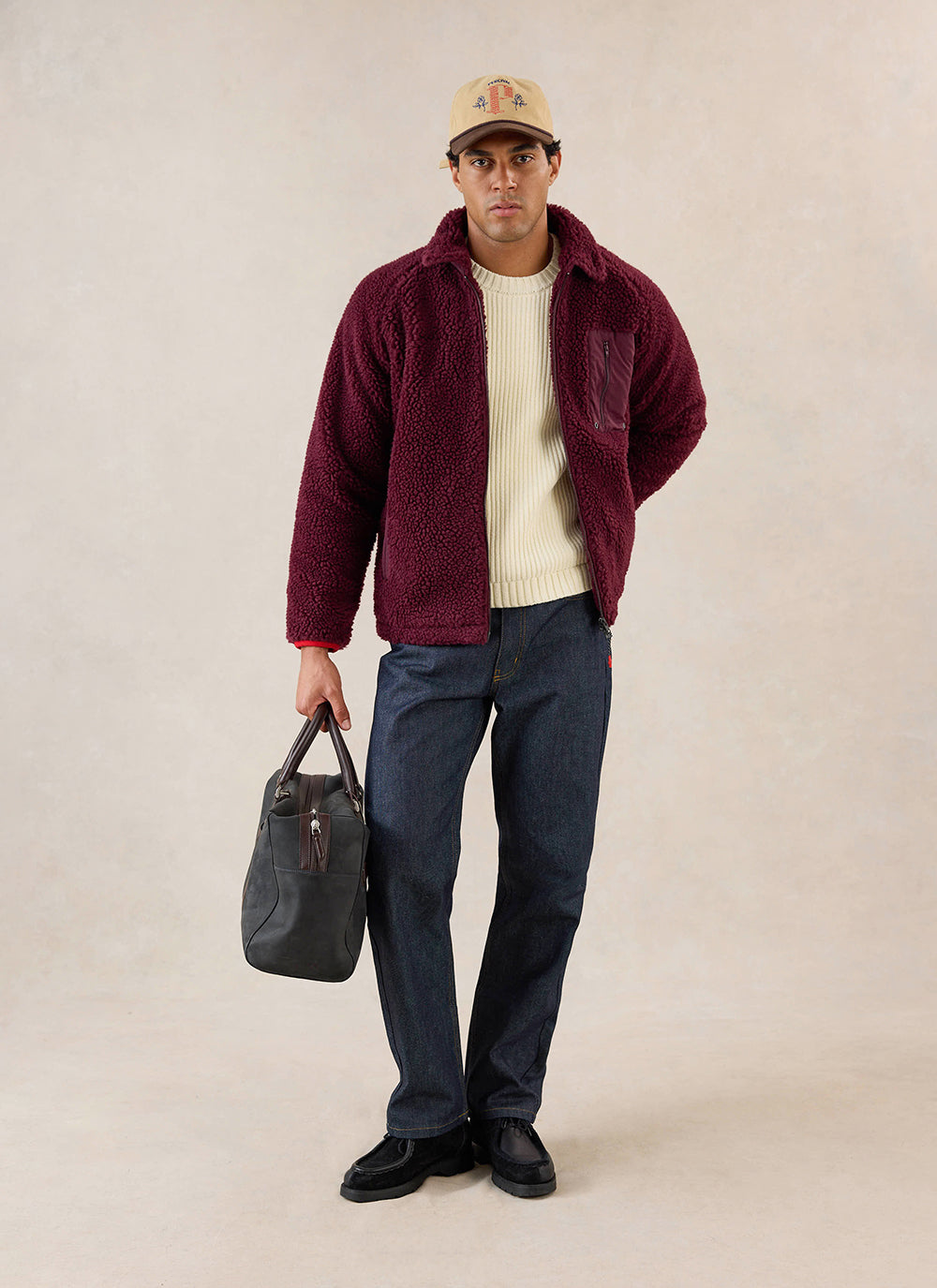 A model wears the merlot Percival wool fleece jacket over a cream knit jumper, paired with dark jeans and holding a bag.