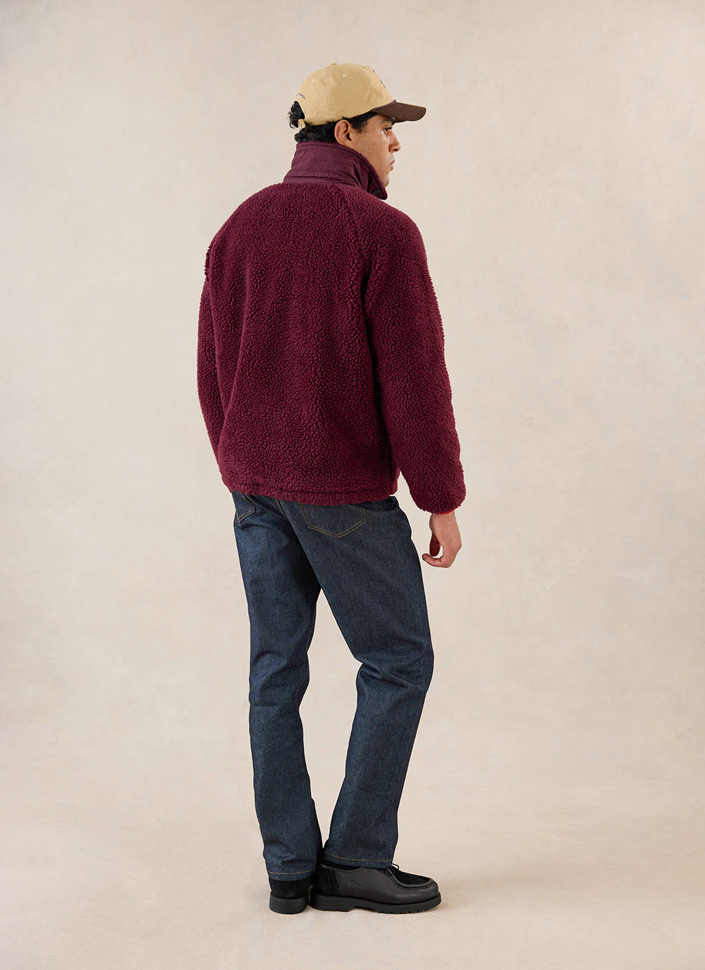 A rear-view shot of a model wearing the merlot Percival fleece jacket, styled with dark denim jeans and a cap.