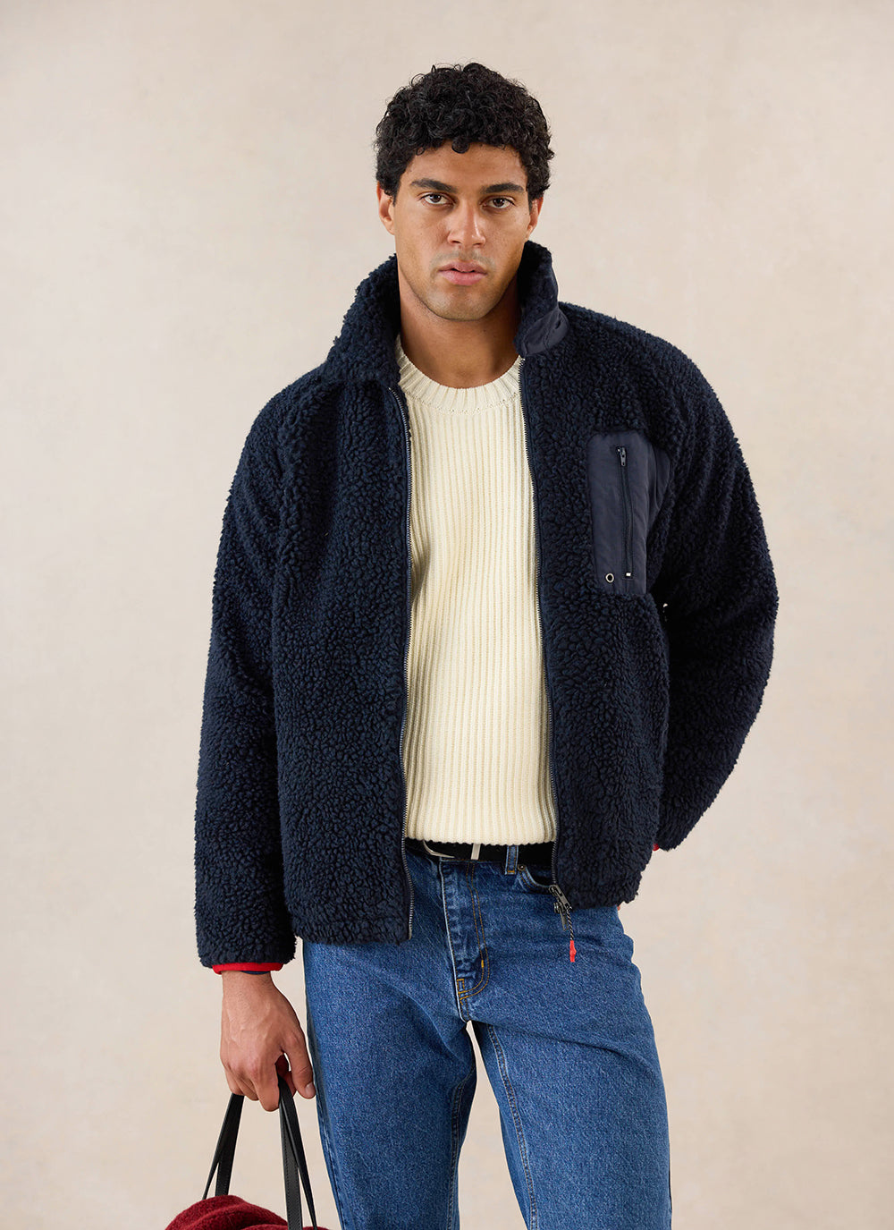 A model wears the navy fleece jacket from Percival with a cream jumper and blue jeans in a studio setting.