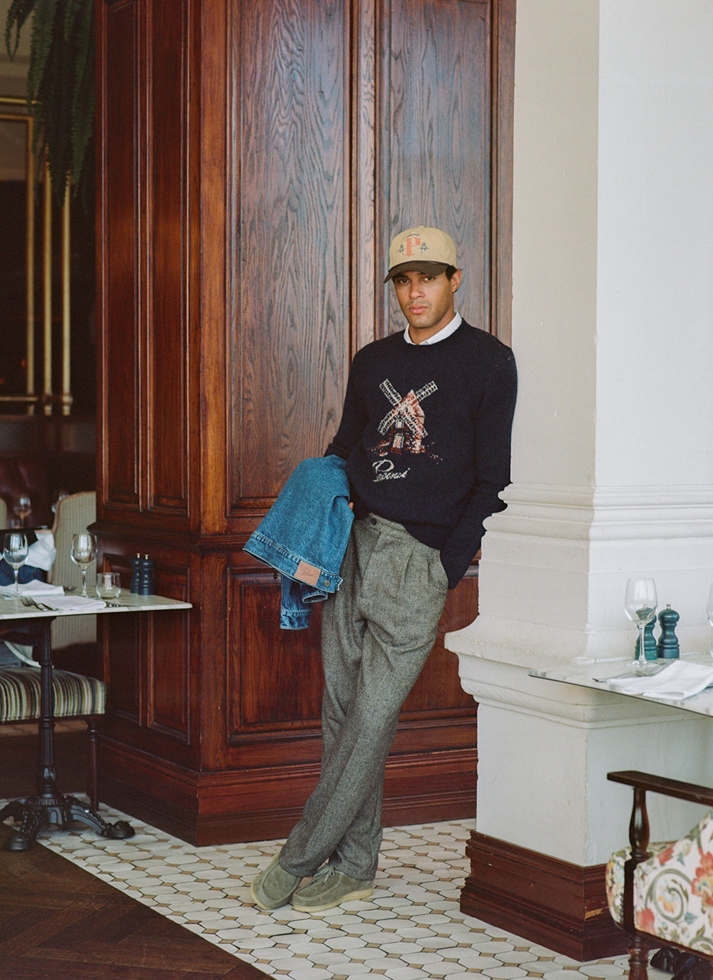 Model in navy Percival Miller jacquard jumper with windmill graphic, holding denim jacket, with grey trousers and cap.