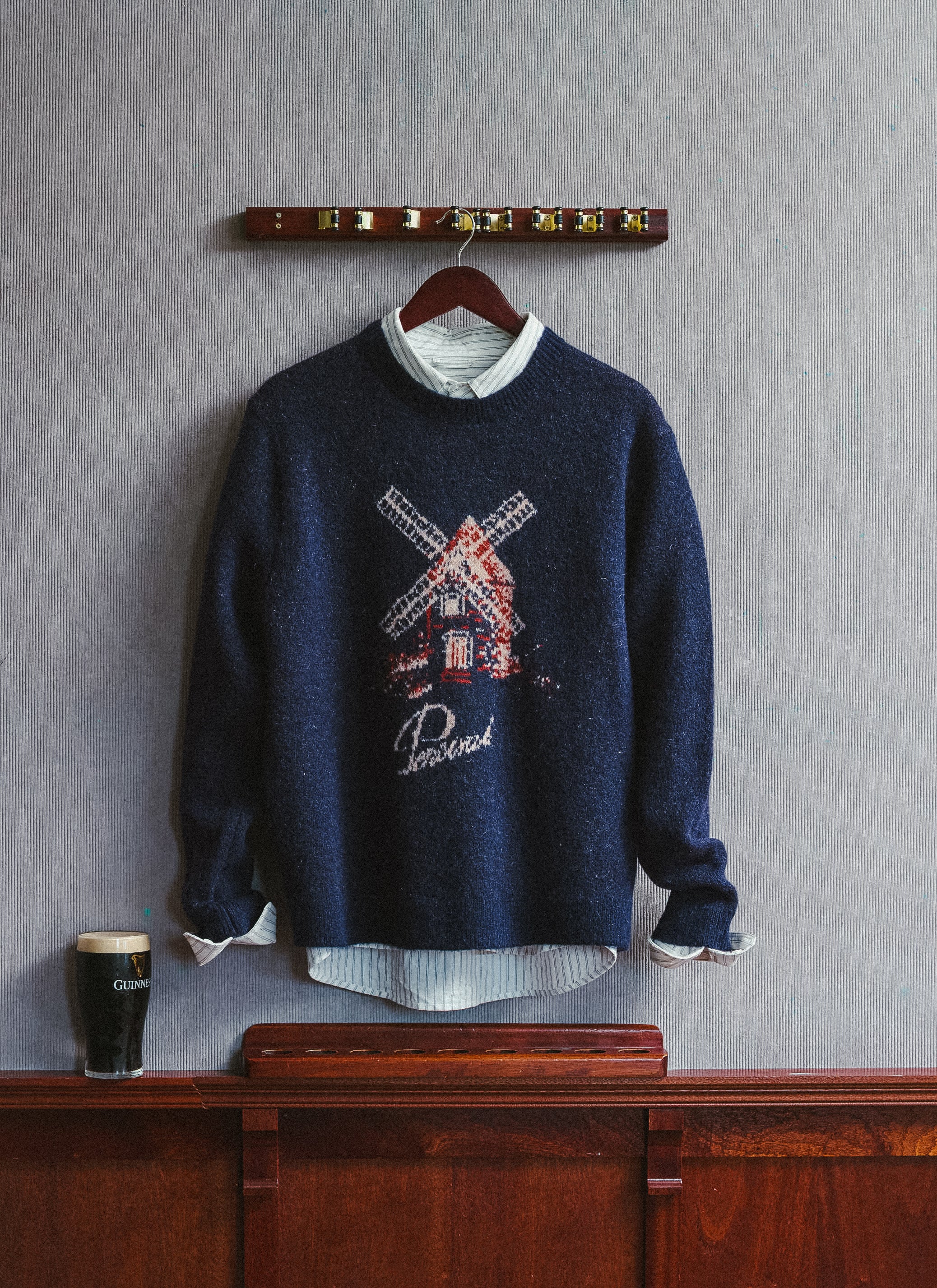 Percival navy Miller jacquard jumper layered over a striped shirt, hanging on a wooden hook with a pint nearby.