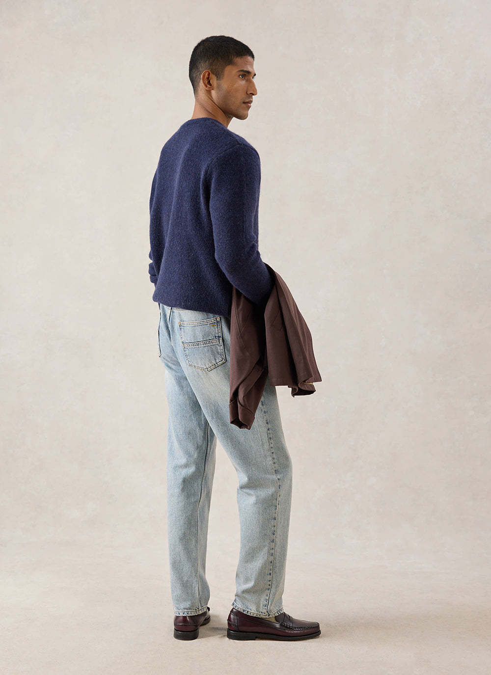 A model wears the navy Percival Miller Boxy Jacquard Jumper from behind with light-wash jeans and a jacket.