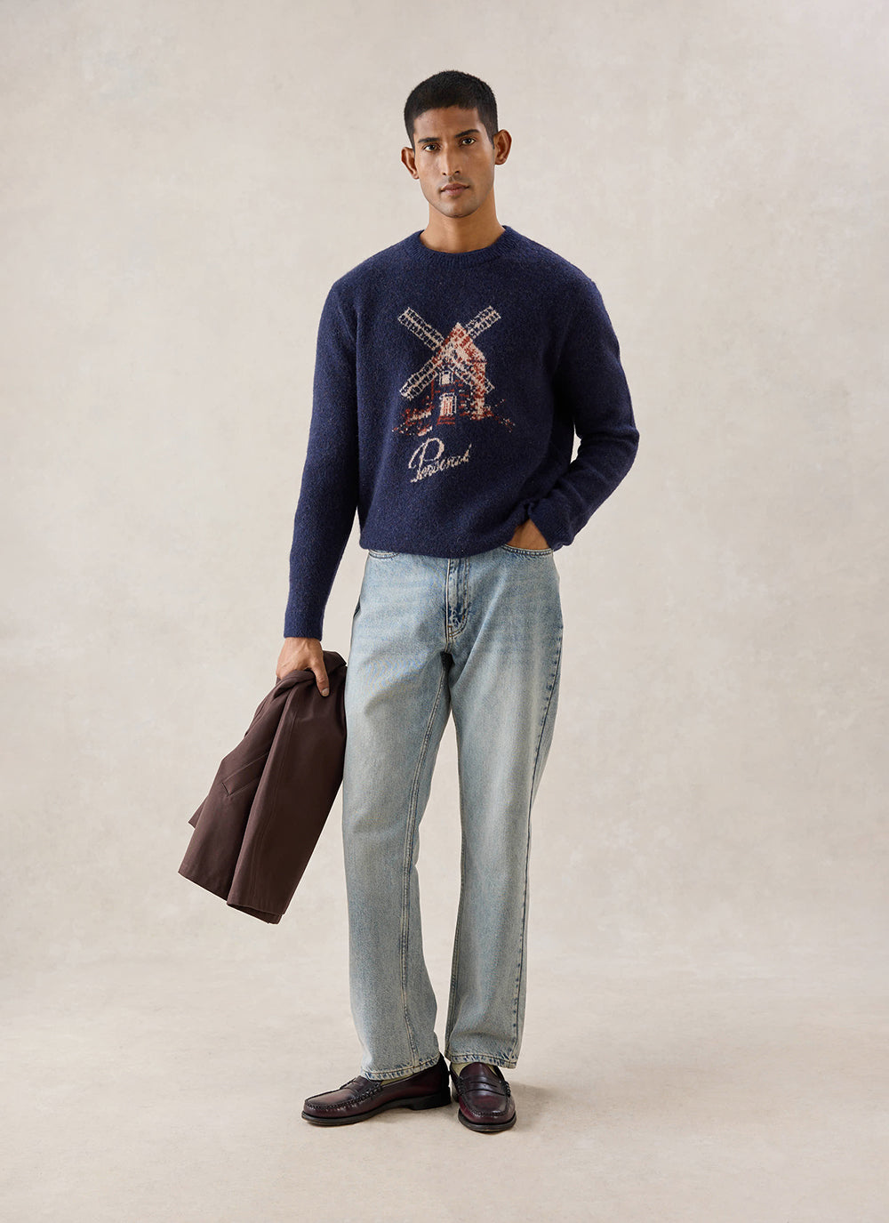 A model wears the navy Percival Miller Jacquard Jumper with a windmill graphic, styled with light blue jeans and loafers.