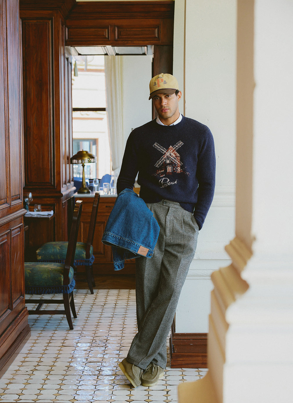 A model wears the Percival Miller navy jumper, birdseye trousers, and beige Tucker cap in a restaurant interior.