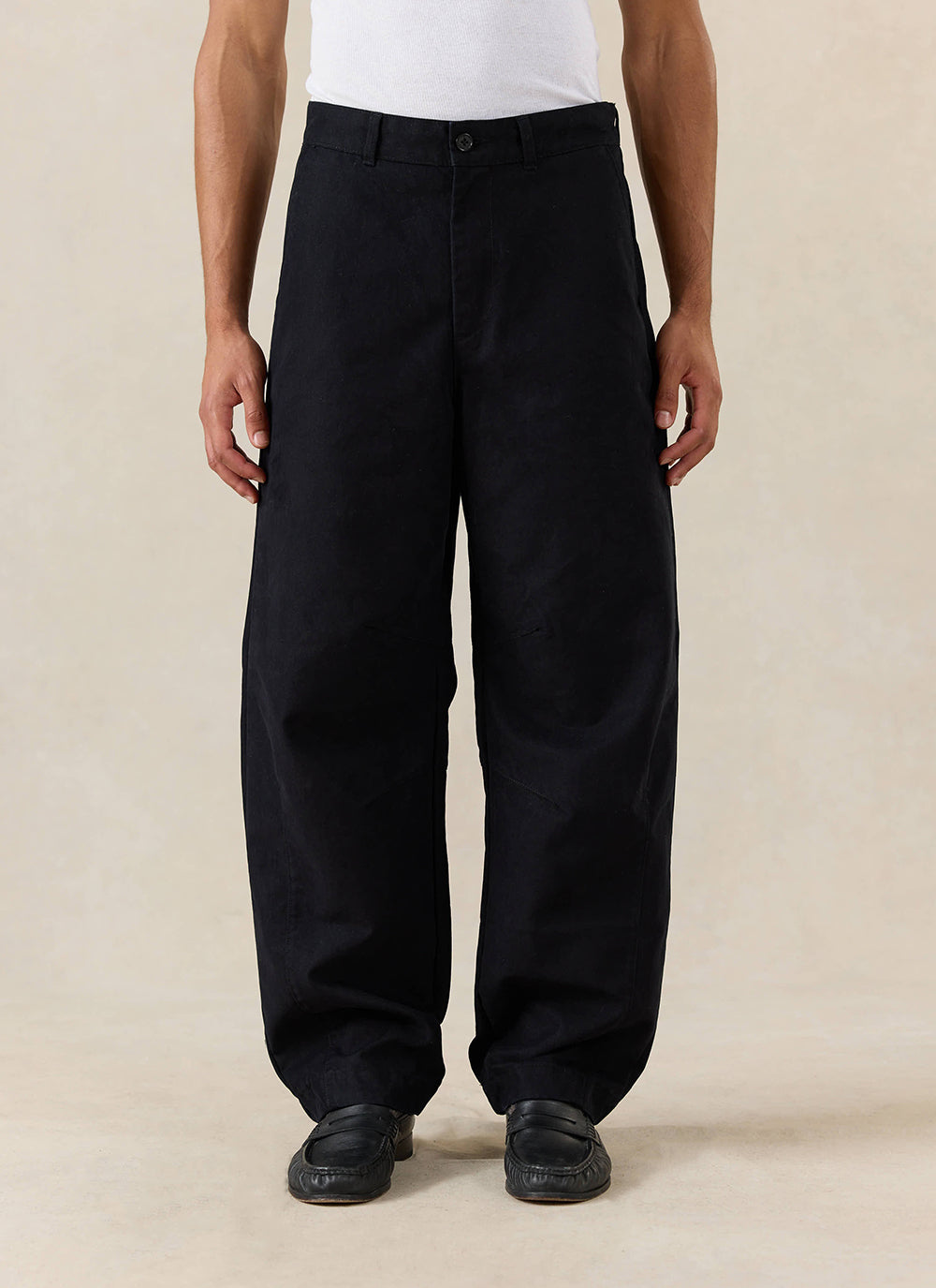 A model wears the black Percival Milo Relaxed Barrel Leg Trousers, styled with a white vest and black loafers.